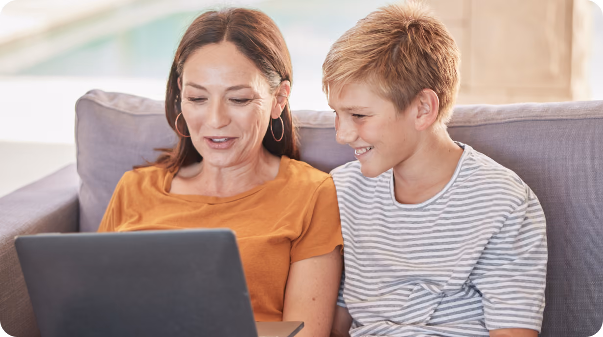 A woman and a smiling boy sit together on a couch looking at a laptop.