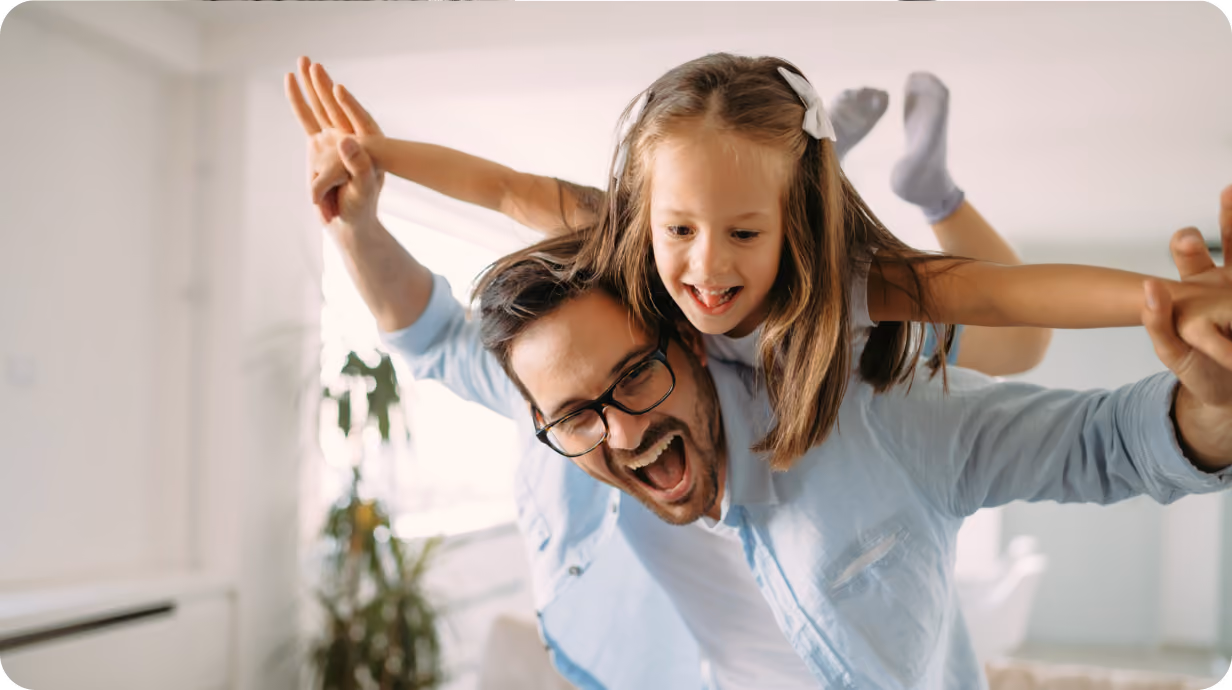 A laughing man carries a happy young girl on his shoulders indoors, both with arms outstretched.