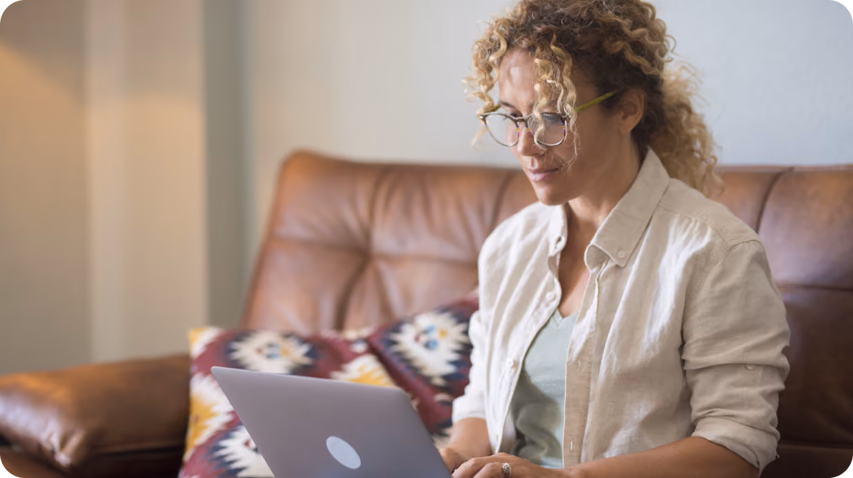 A woman with curly hair and glasses sits on a leather couch working on a laptop.