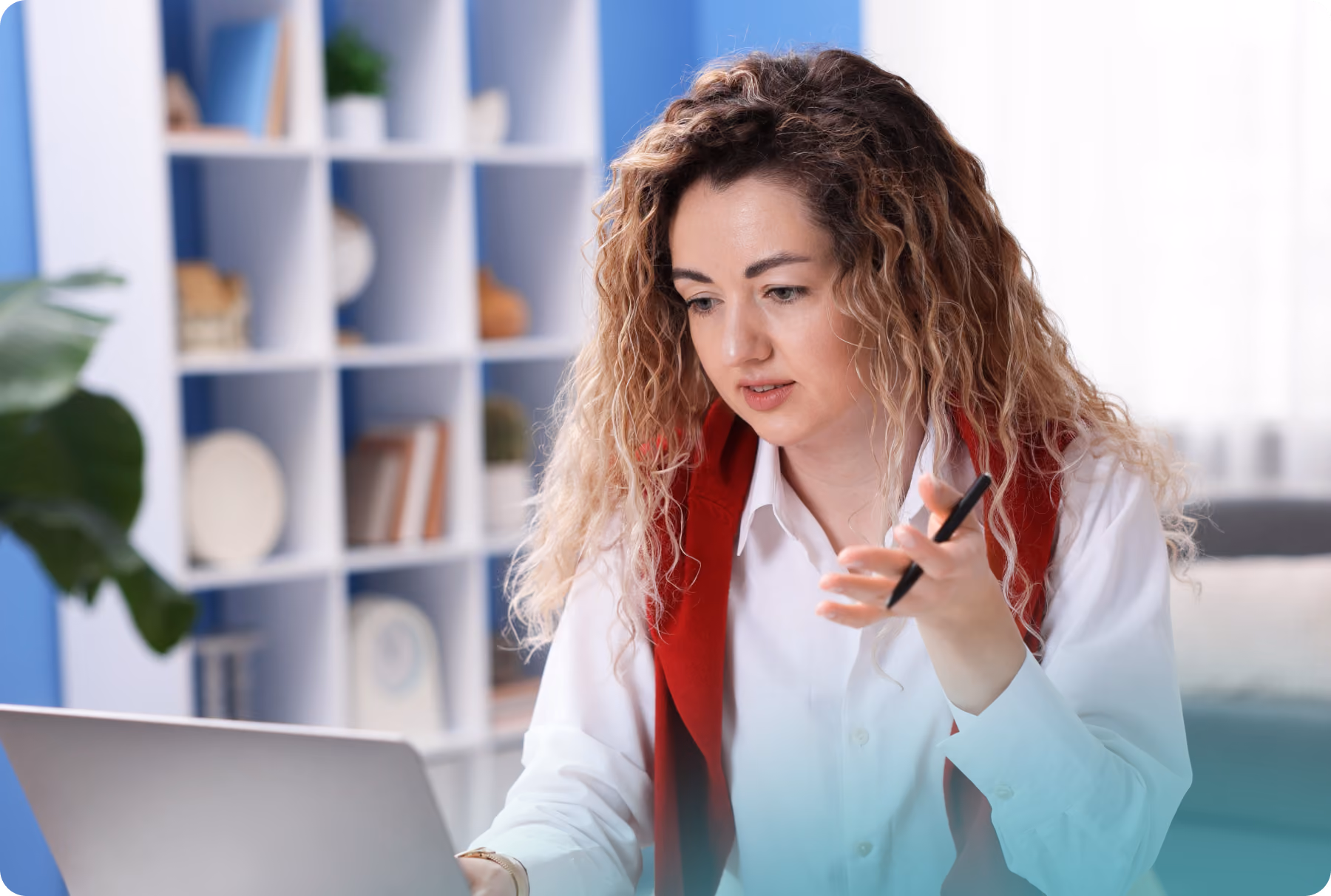 A woman with curly hair, wearing a white shirt with a red sweater over her shoulders, holds a pen and looks at a laptop while speaking.