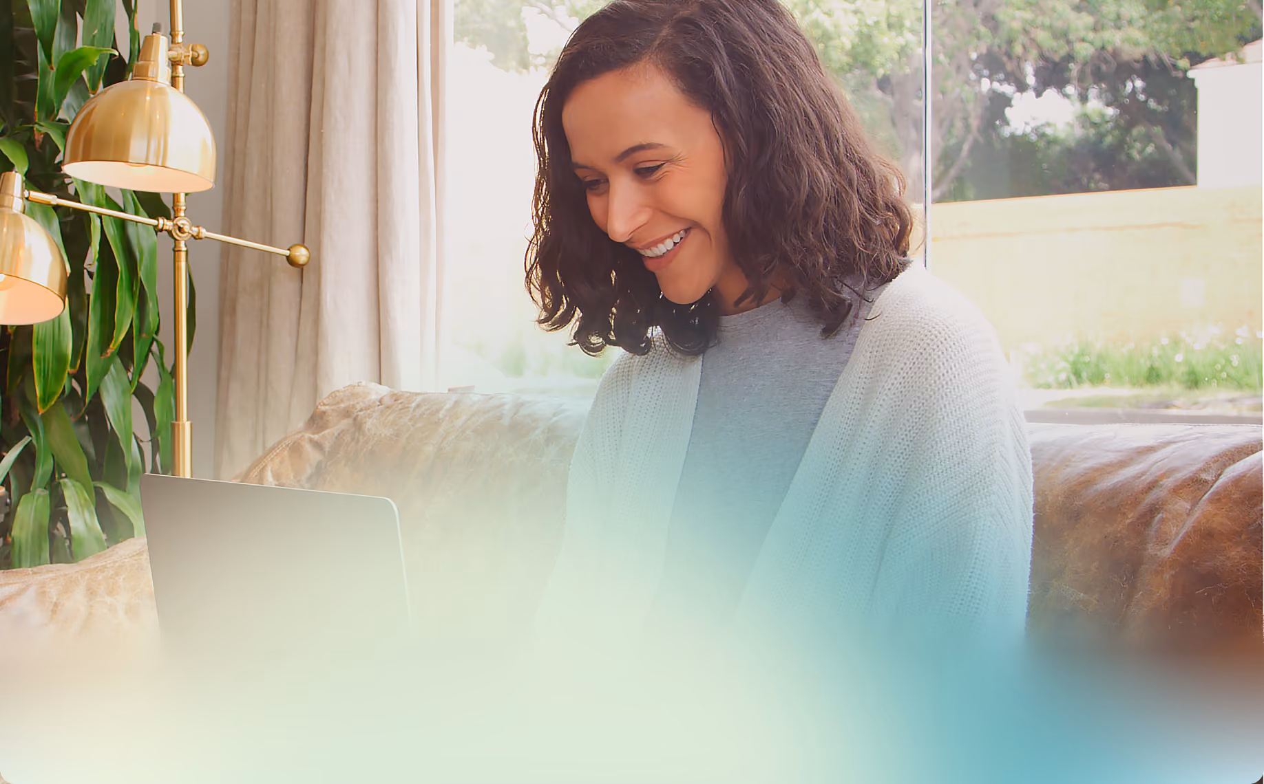 A smiling woman with curly dark hair sits on a couch by a window, looking down at a laptop.