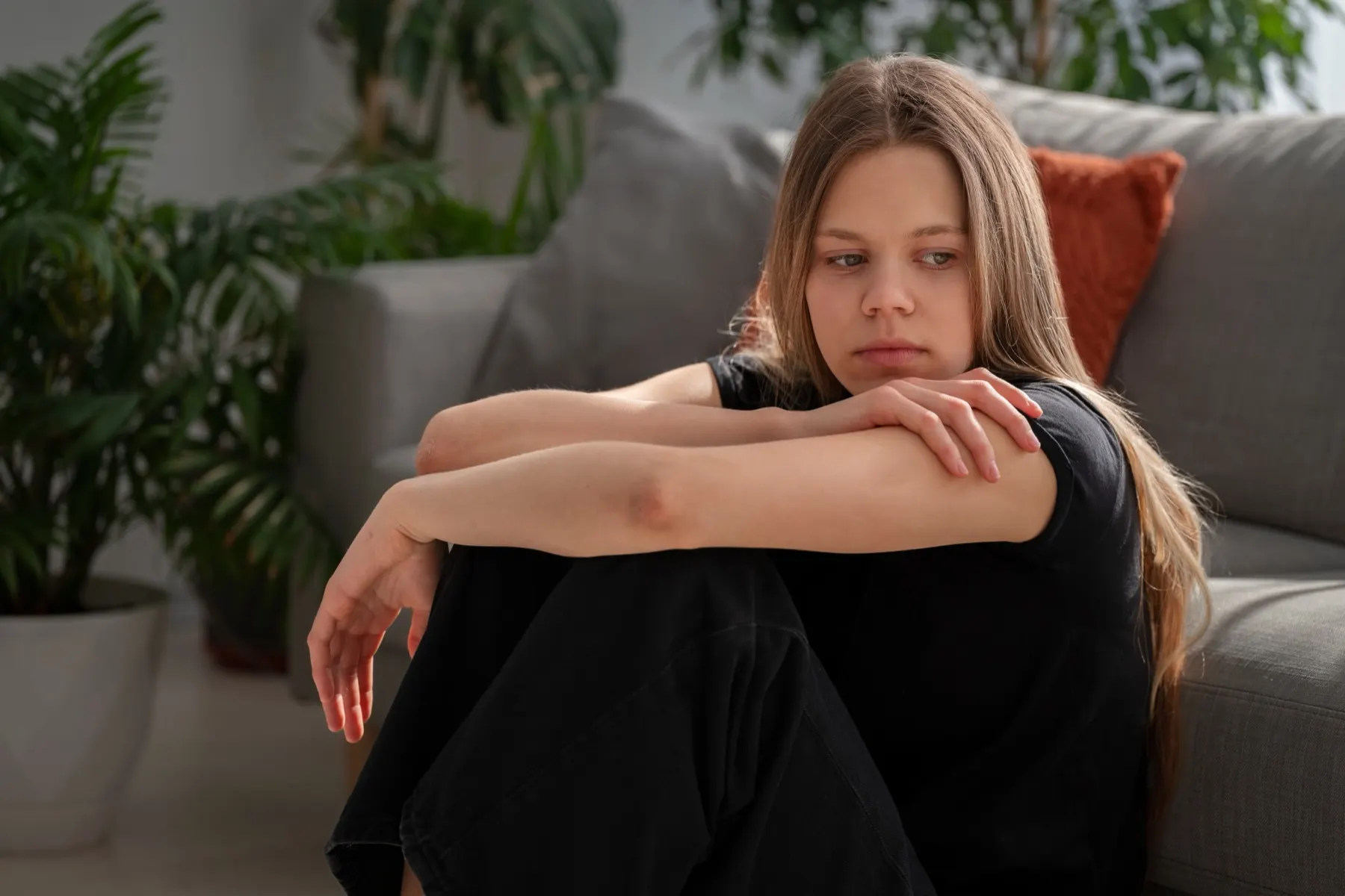 Teen girl sitting on the floor by a couch, hugging her knees and looking worried