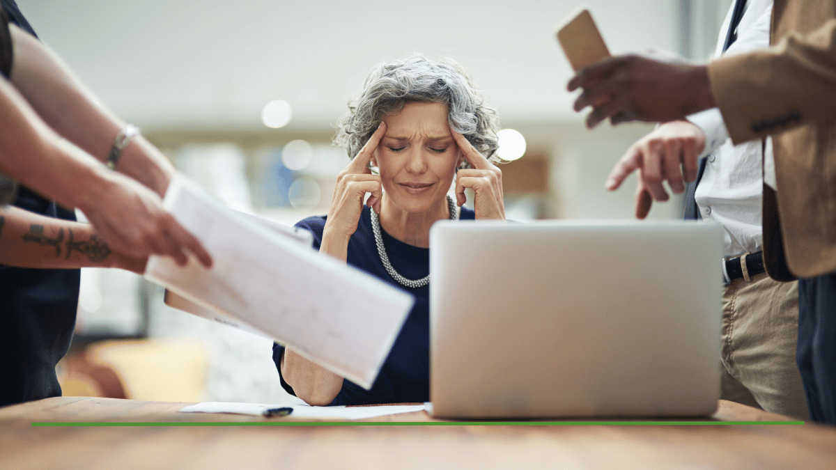 Stressed woman rubbing her temples at a laptop while a colleague hands her paperwork, illustrating the confusion around Roth IRA rules and downsides