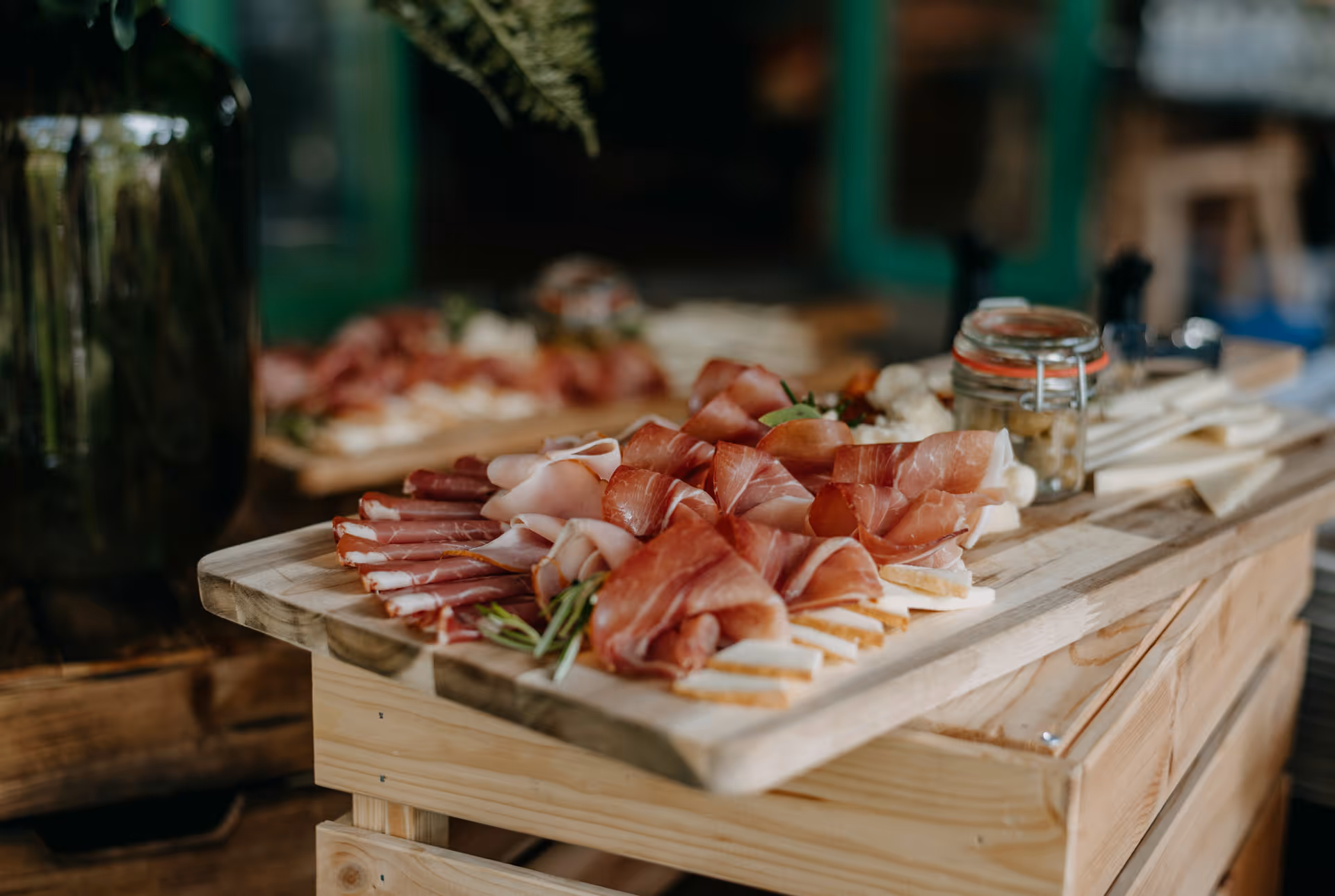 Wooden board with an assortment of thinly sliced cured meats, cheese, herbs, and a jar of olives.
