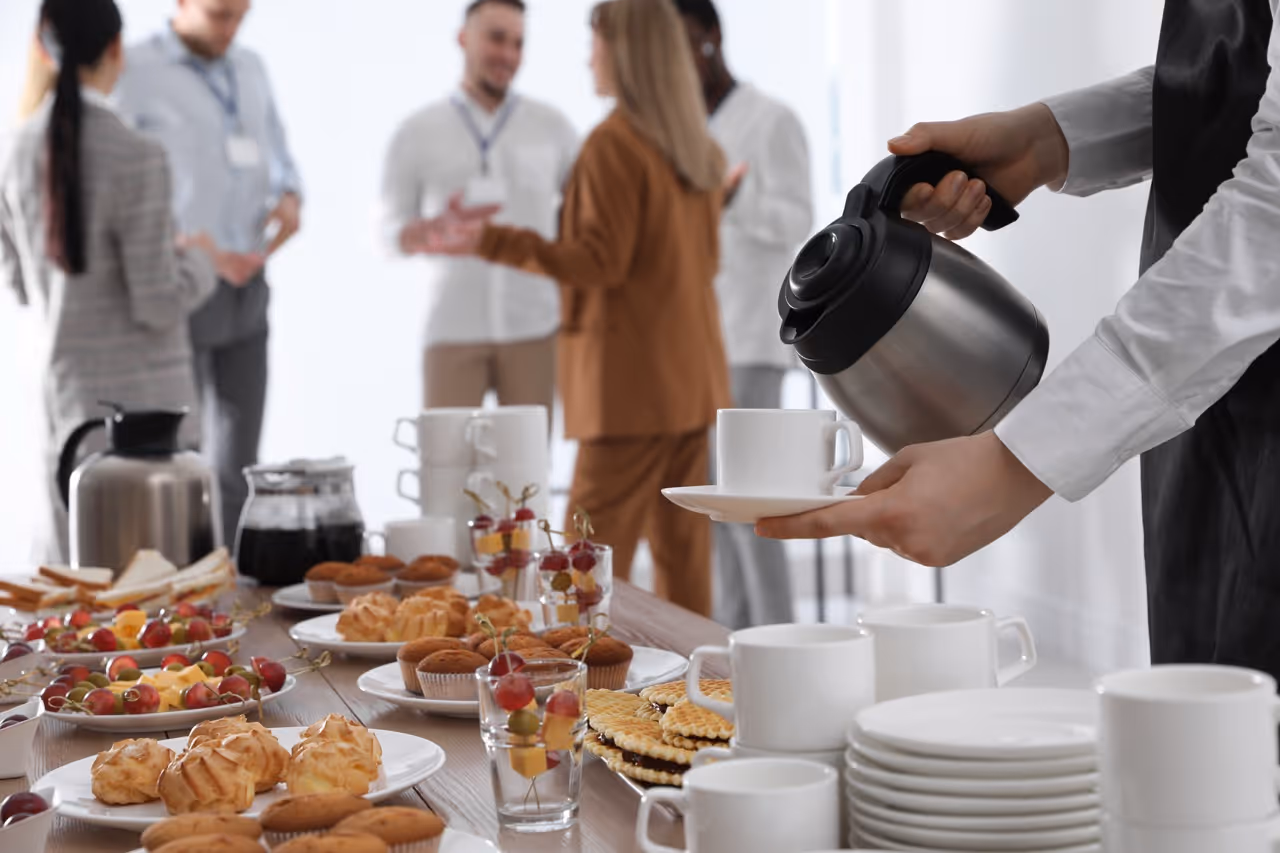 Person pouring coffee into a white cup at a table with assorted pastries and fruits, with people conversing in the background.