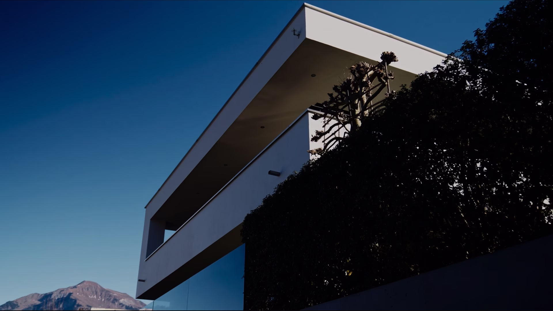 Modern white building with large balcony and glass railing against a clear blue sky and mountain background.