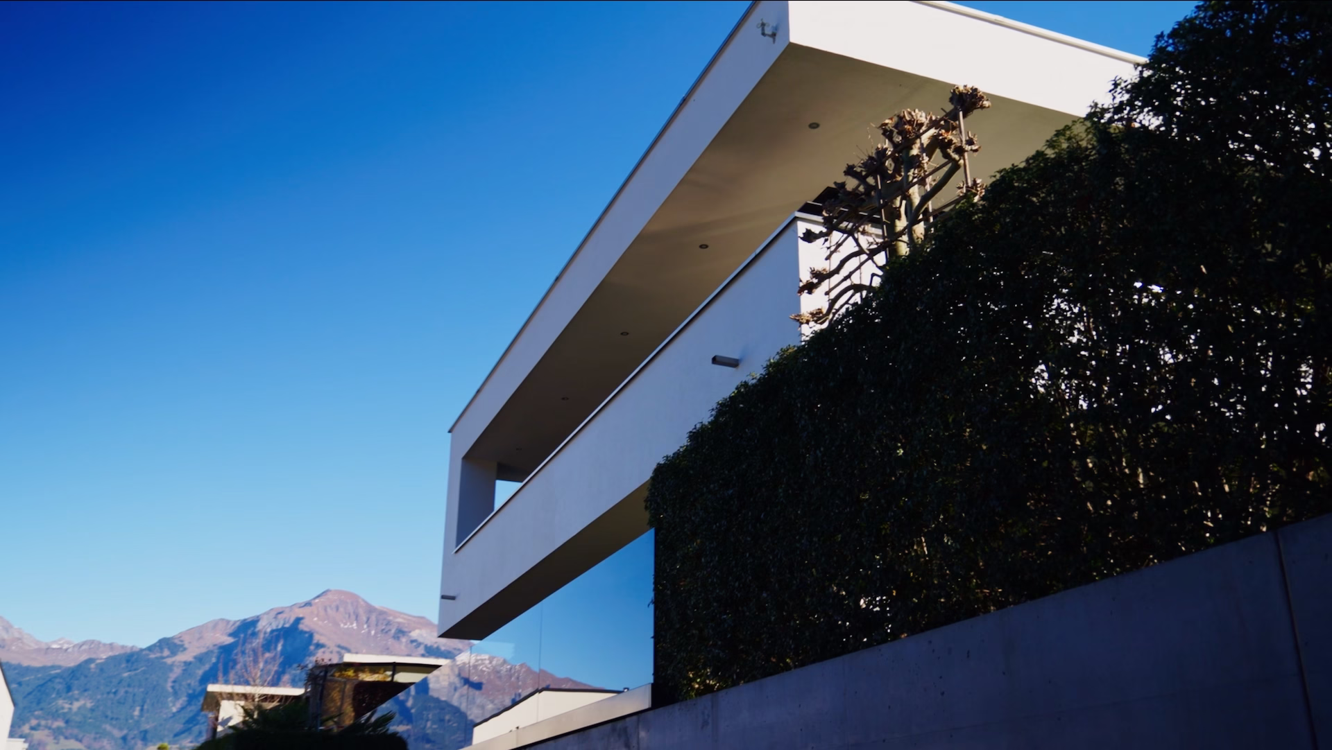 Modern white multi-story house with large balcony and hedge in front, set against clear blue sky and mountain backdrop.