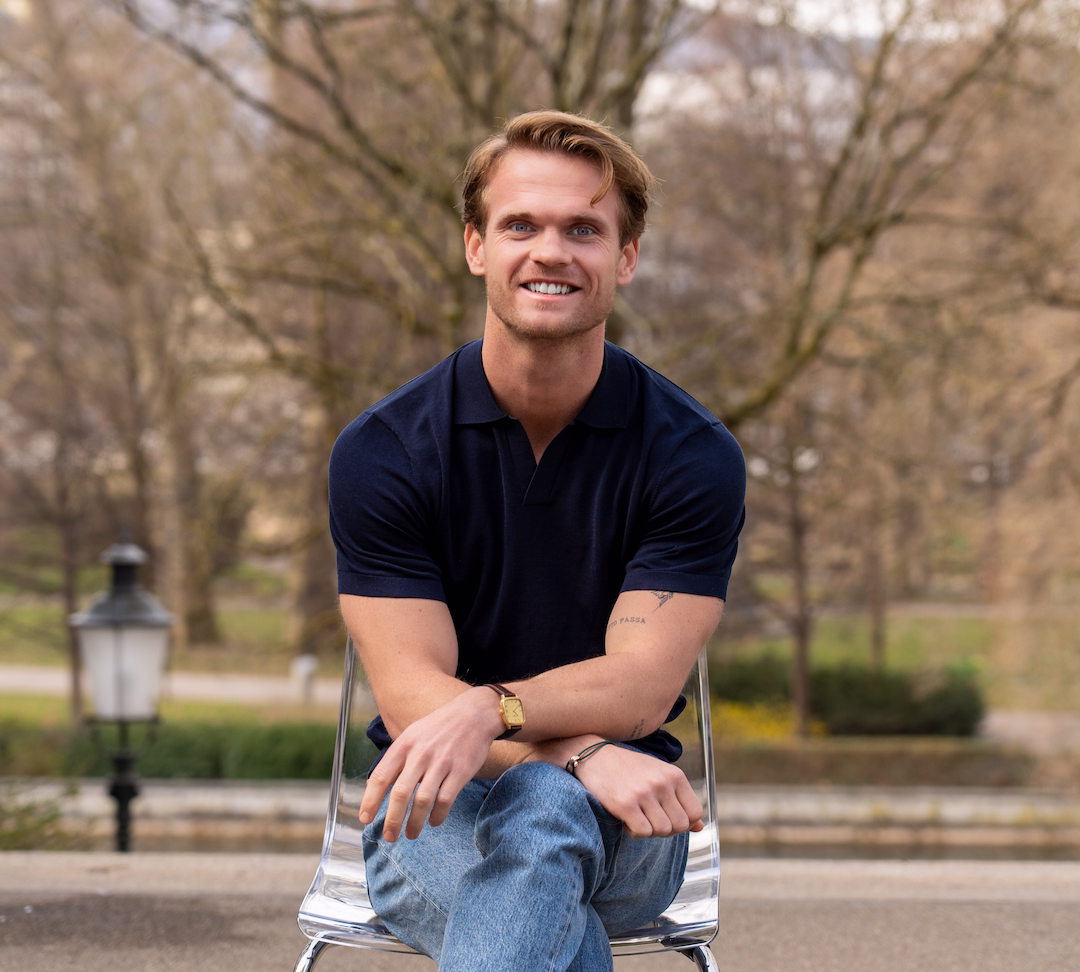 Smiling young man with light brown hair wearing a navy polo shirt and jeans sitting cross-legged on a transparent chair outdoors with blurred trees in the background.