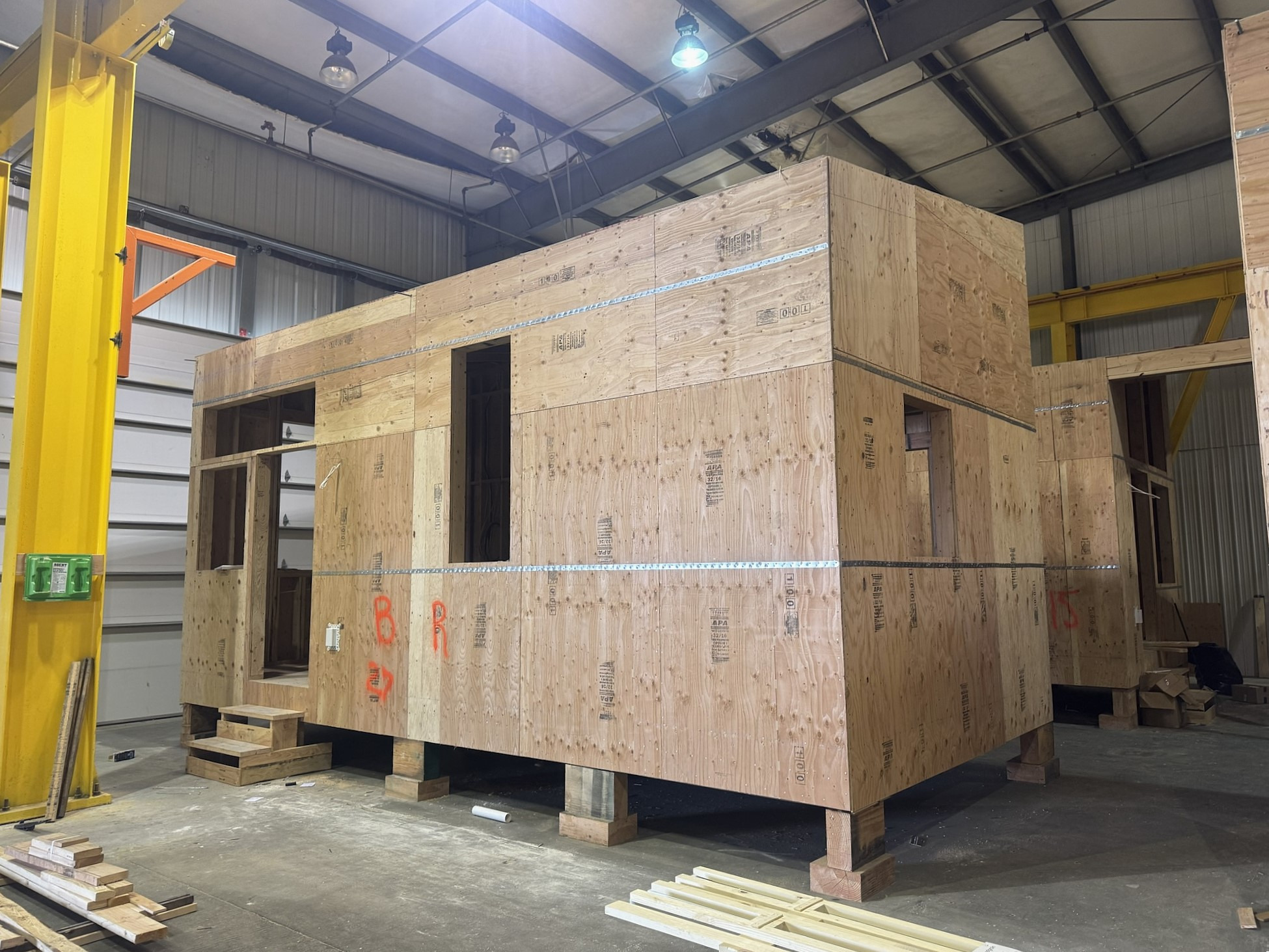 A tiny home module framed and sheathed on cribbing inside the fabrication shop, under a yellow gantry crane
