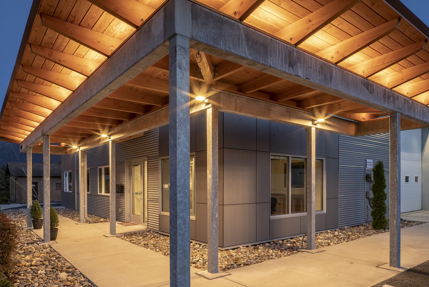 Lit timber canopy and galvanized steel colonnade wrapping the entry of Dawson's Juneau headquarters at dusk