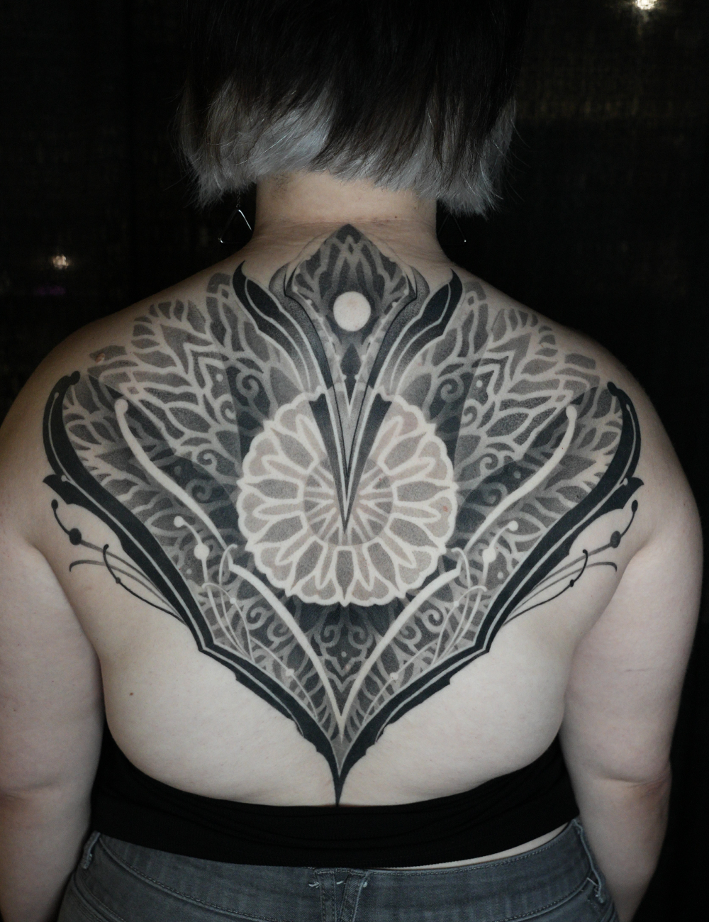 Ornamental upper back tattoo of a symmetrical mandala with bold black shapes by Vanessa