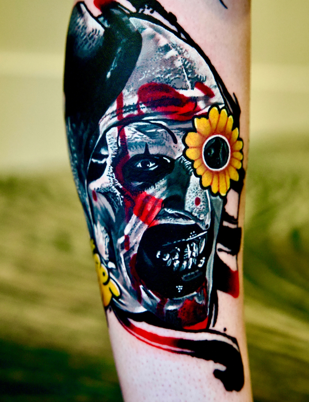 Realism forearm tattoo of a horror clown face with red splashes and a sunflower by Juanky