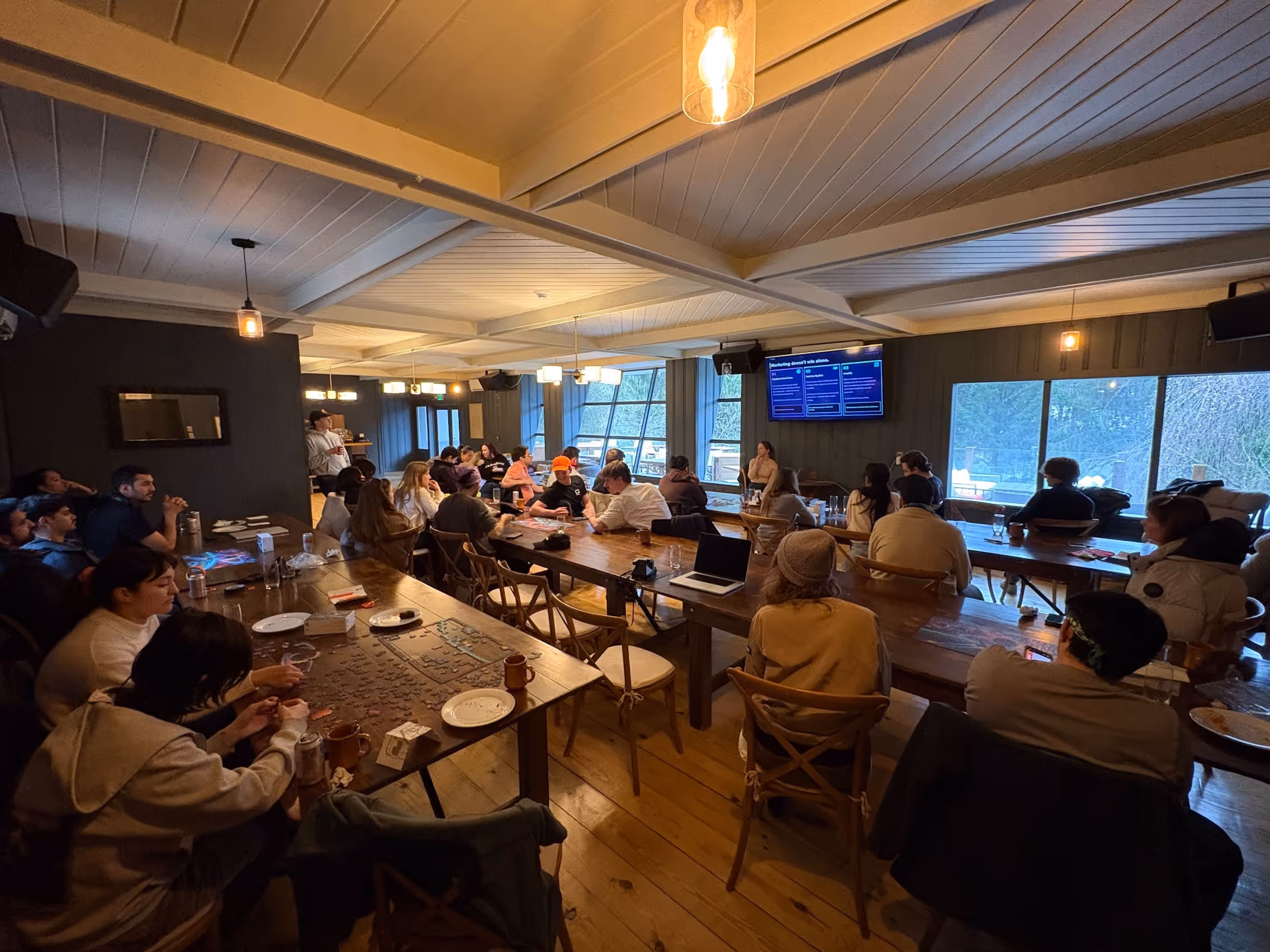 Group of people seated around wooden tables in a room, watching a speaker near a screen.
