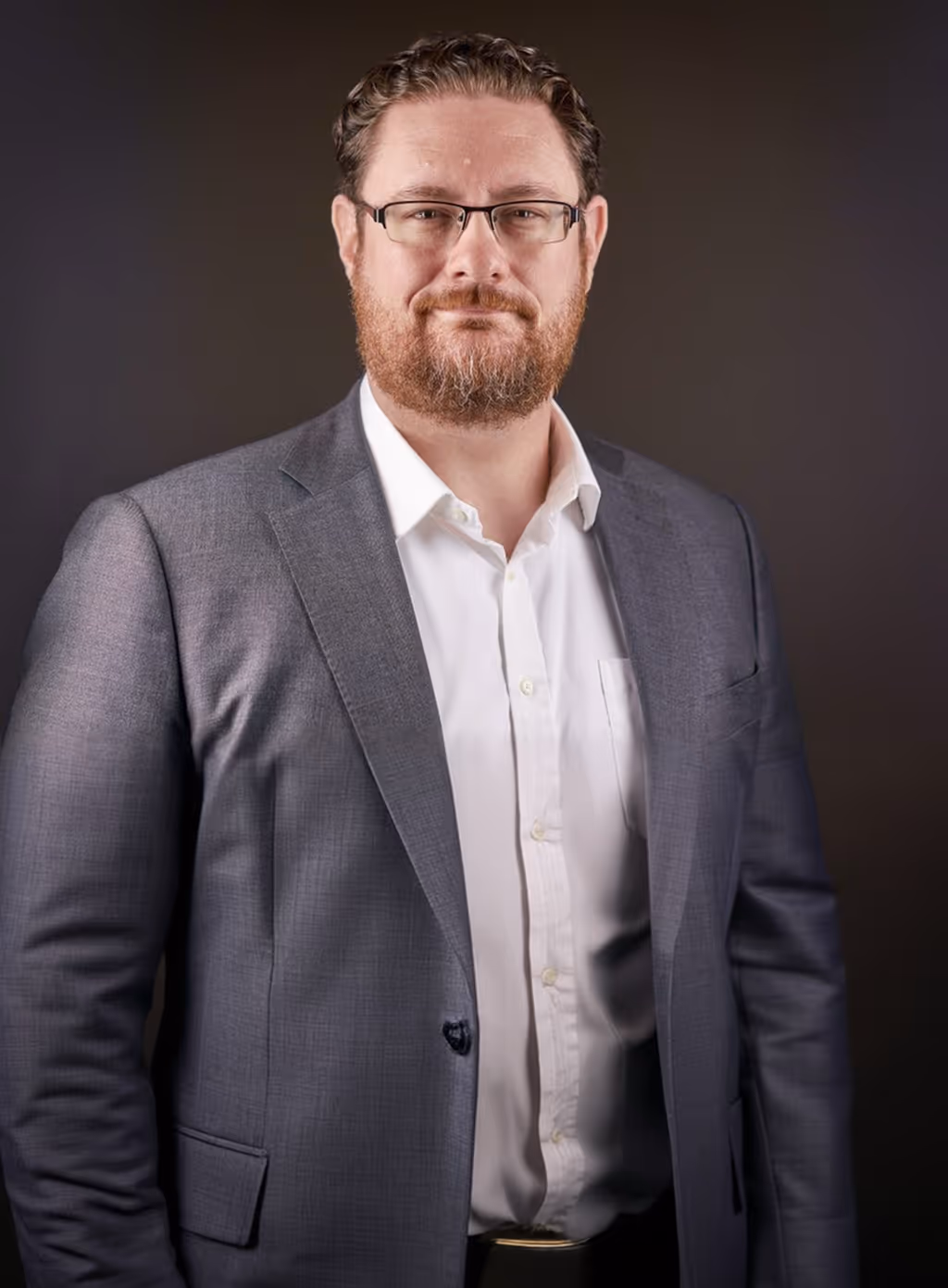 Man with glasses, beard, and curly hair in a gray suit jacket and white shirt against a dark background.