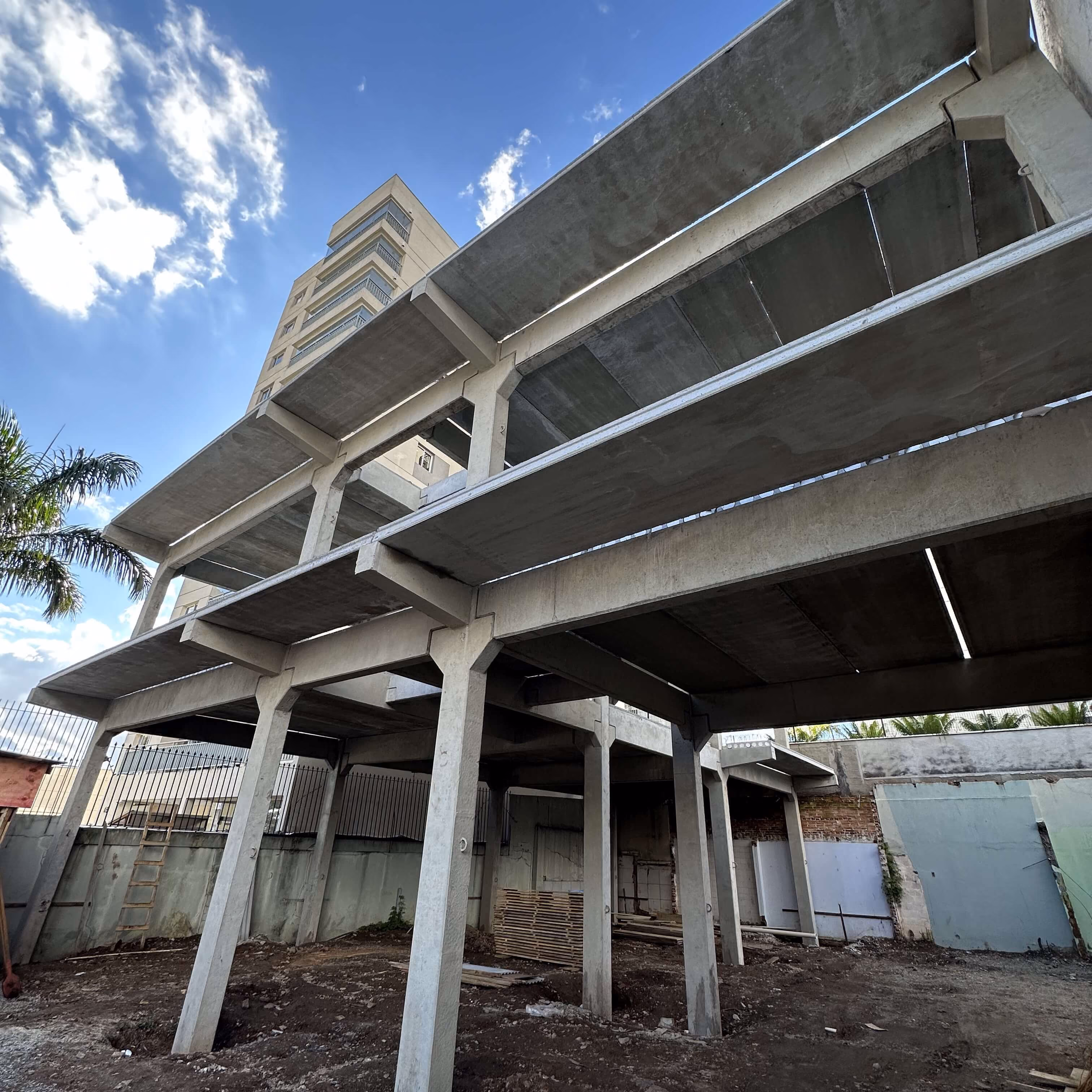 Estrutura de concreto em construção vista de baixo, com céu azul e edifício ao fundo.