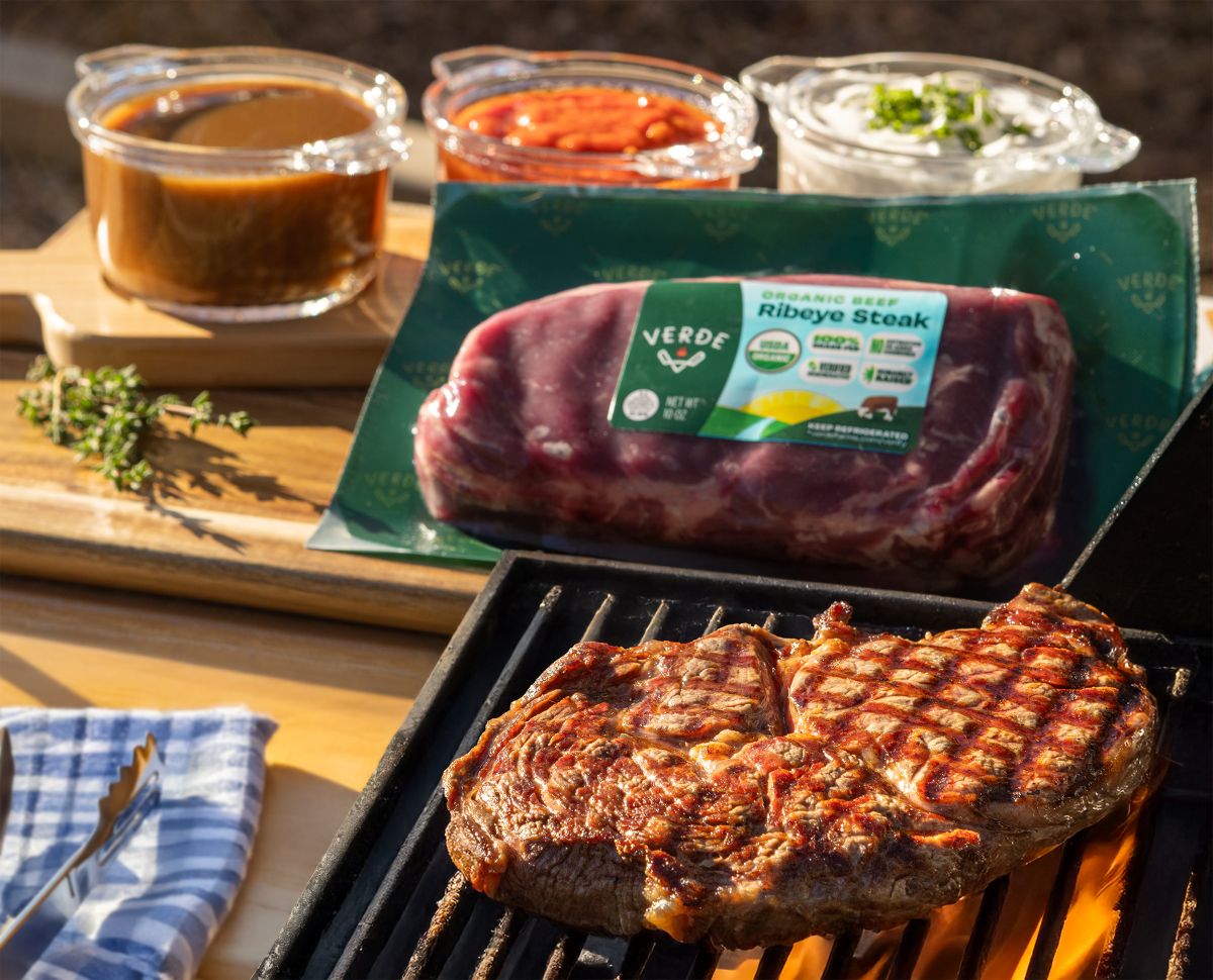 Grilled ribeye steak cooking on a grill with raw ribeye and sauces on a wooden board nearby.