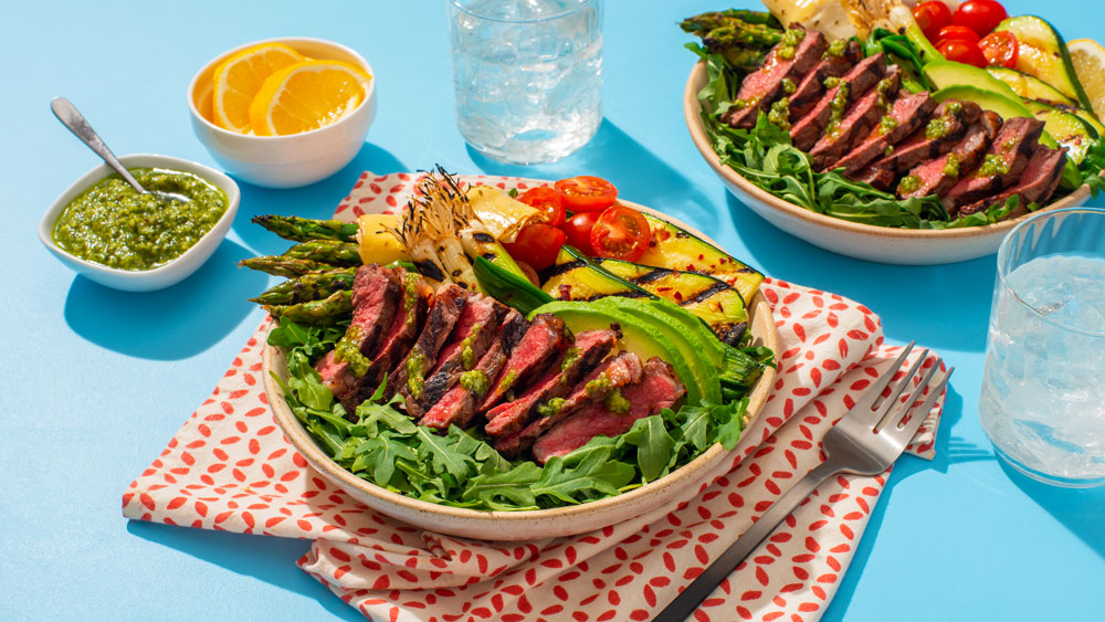 Two bowls with grilled steak, asparagus, avocado, cherry tomatoes, and greens, with lemon and pesto sauce on the side.