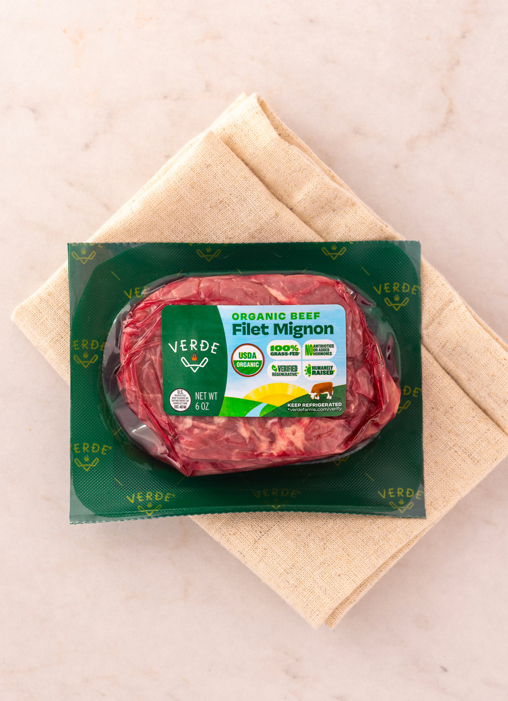 Vacuum-sealed package of organic filet mignon beef from Verde Farms on a beige cloth napkin with peeled potatoes and a sprig of rosemary nearby.
