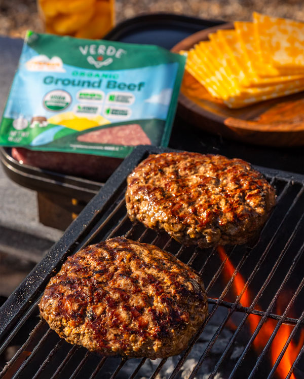 Grilled ribeye steak with grill marks cooking on a barbecue with packaged raw ribeye steak and three sauces in clear containers on a wooden board nearby.