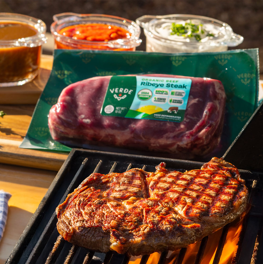 Grilled ribeye steak with grill marks cooking on a barbecue with packaged raw ribeye steak and three sauces in clear containers on a wooden board nearby.