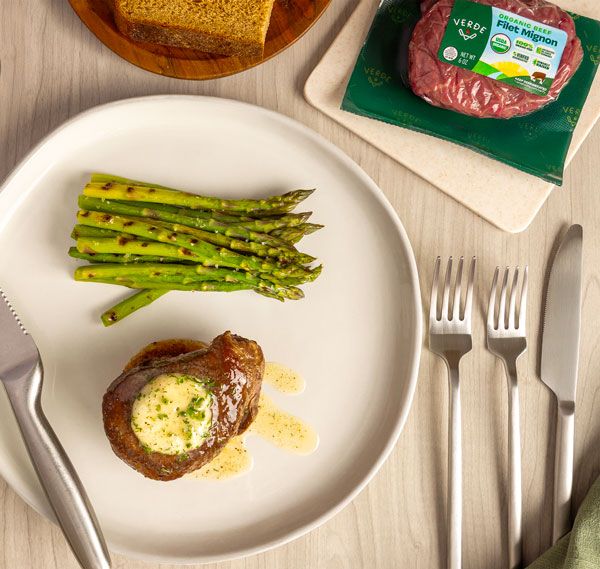 Grilled ribeye steak with grill marks cooking on a barbecue with packaged raw ribeye steak and three sauces in clear containers on a wooden board nearby.