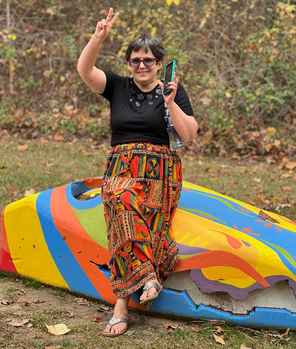 An image of a woman with a disability standing outside smiling, one her phone, and giving a peace sign