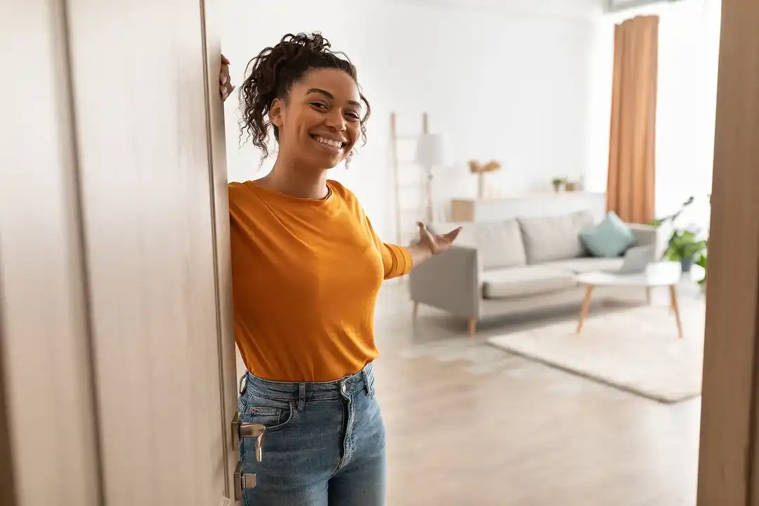 A young woman opening the front door and extending her arm to invite the viewer into her home