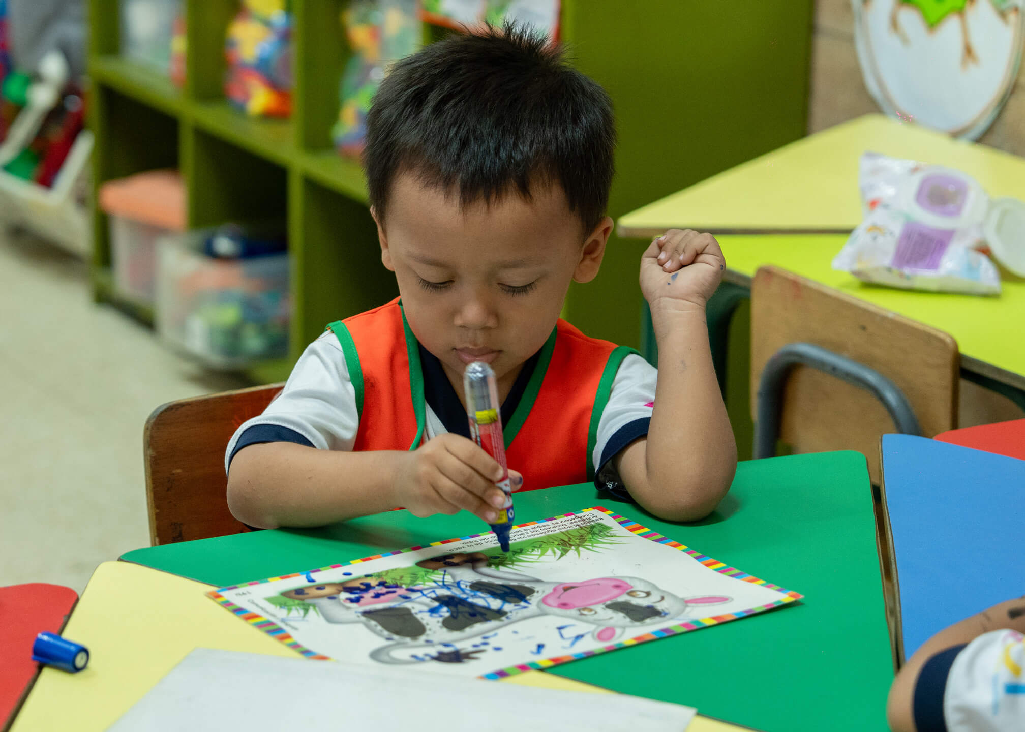 Niño pequeño sentado en una mesa de colores en un aula, coloreando con un marcador azul un dibujo de una vaca en una hoja.