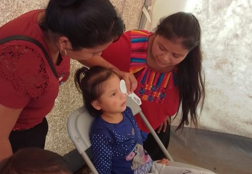 Una niña pequeña sentada en una silla mientras dos mujeres le miden la temperatura con un termómetro infrarrojo en la frente dentro de un lugar cerrado.