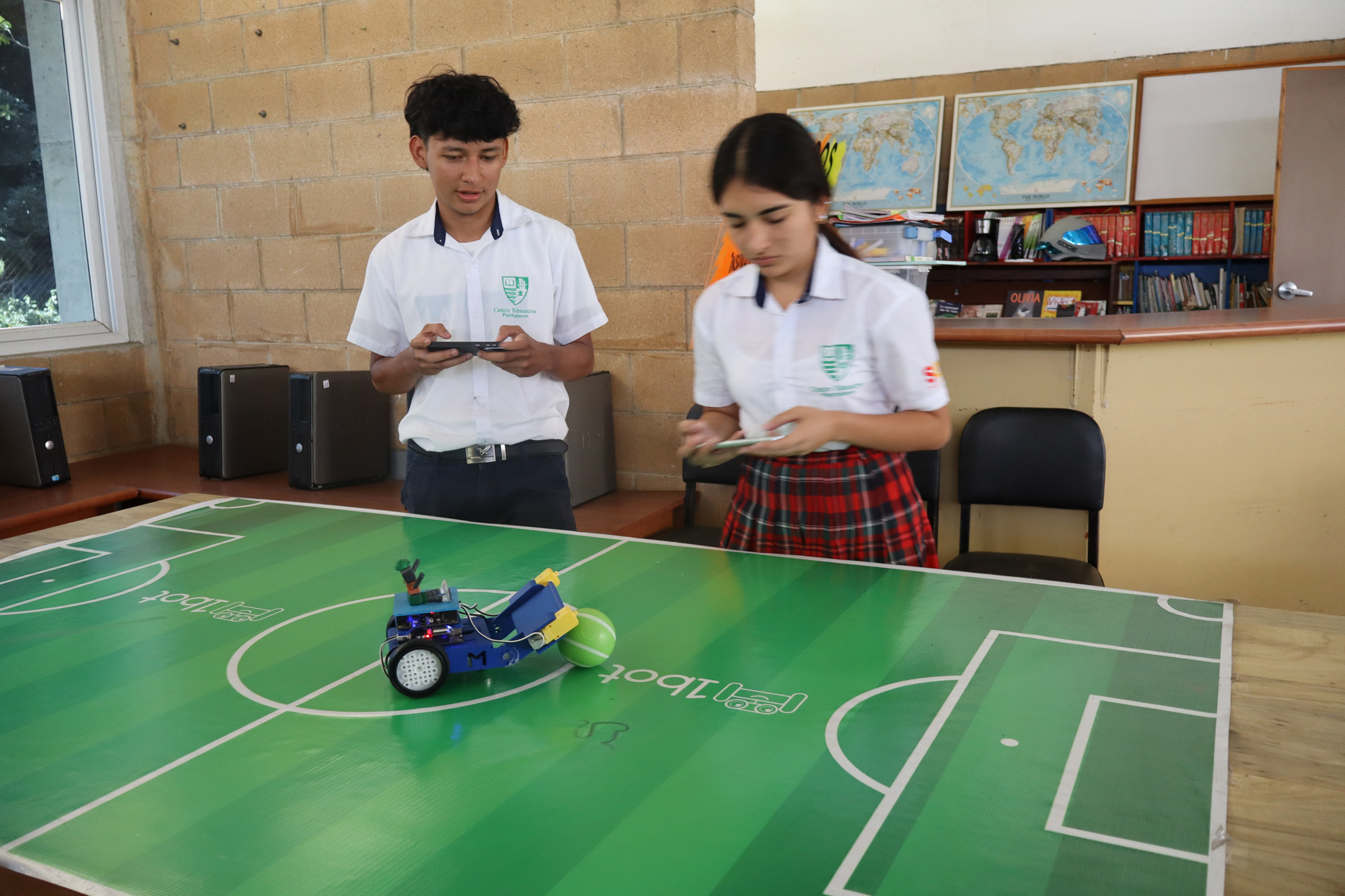 Dos estudiantes en uniformes escolares interactúan con un pequeño robot azul que empuja una pelota verde sobre una mesa con diseño de campo de fútbol, en un aula con computadoras de escritorio y mapas del mundo en la pared.