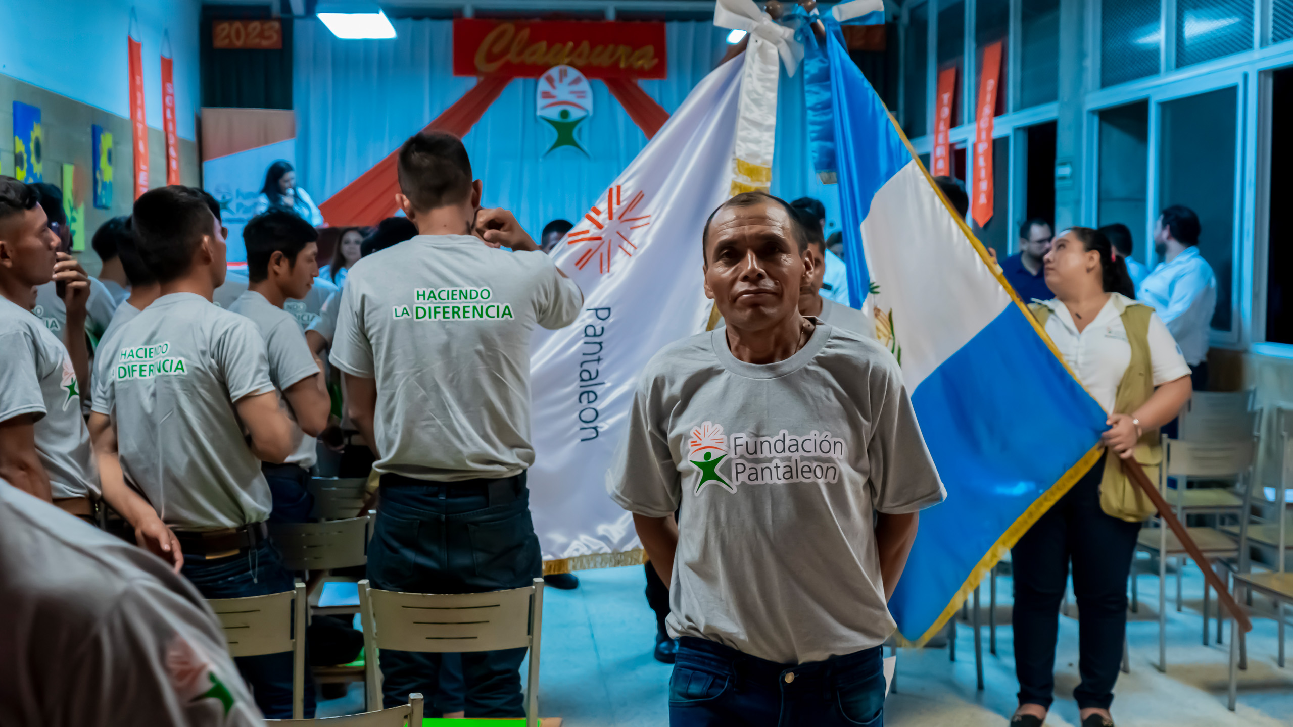 Hombre con camiseta gris que dice Fundación Pantaleón posando frente a un grupo de jóvenes con camisetas que leen Haciendo la diferencia, en un evento con banderas y decoración con la palabra Clausura.