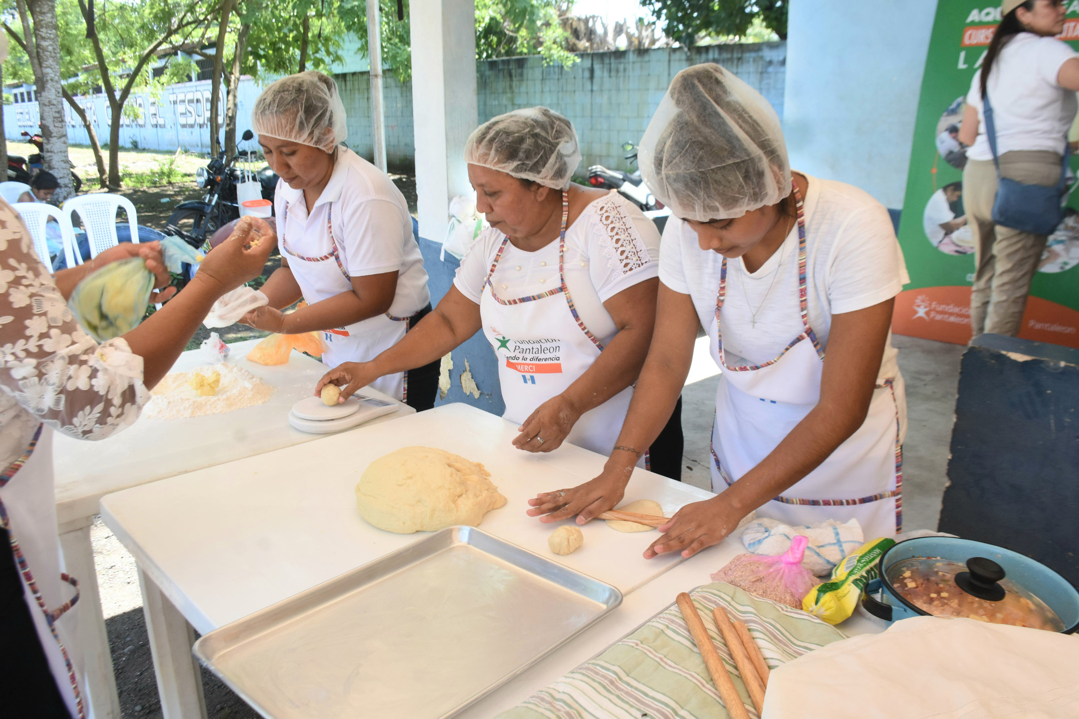 Tres mujeres con redes para el cabello y delantales están preparando masa en una mesa blanca, una enrolla la masa con un rodillo, otra pesa una bola de masa y la tercera ata una bolsa de plástico.