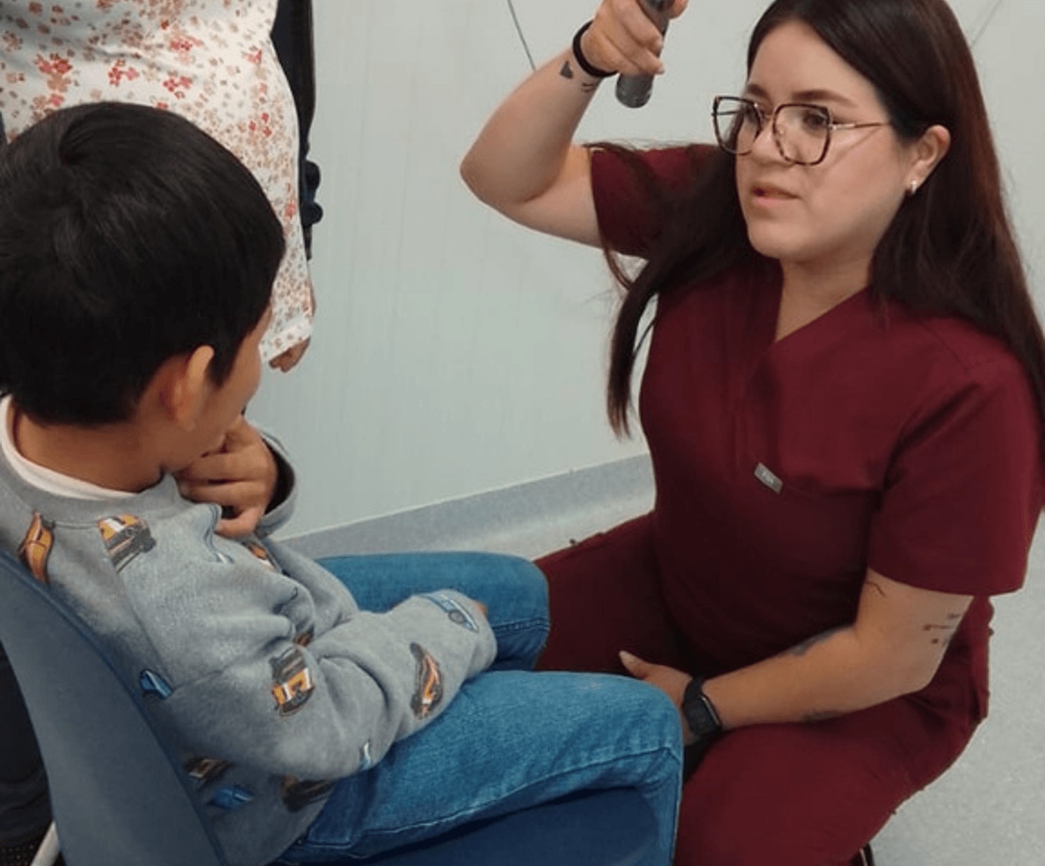 Profesional de la salud con bata color vino y gafas realiza revisión con otoscopio a un niño sentado en una silla azul.