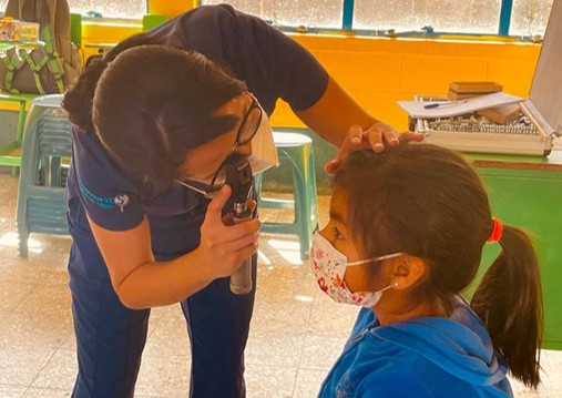 Una profesional de la salud con uniforme azul y mascarilla examina el ojo de una niña que lleva una mascarilla decorada y una sudadera azul, en un ambiente interior con sillas y una mesa al fondo.