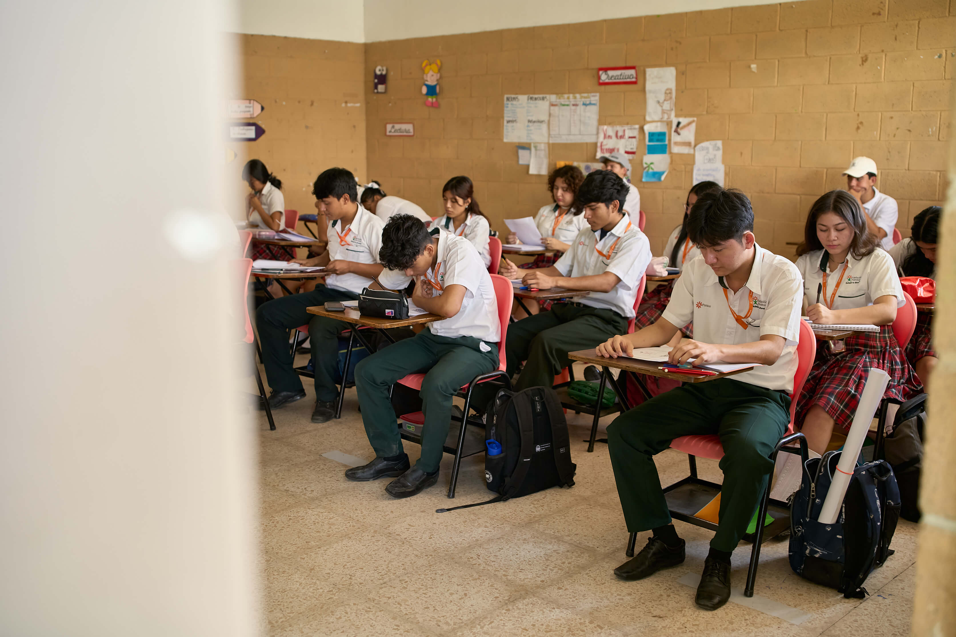 Estudiantes de secundaria en uniforme con camisa blanca y pantalón o falda a cuadros rojos y verdes sentados en escritorios en un aula, concentrados en sus tareas o exámenes.