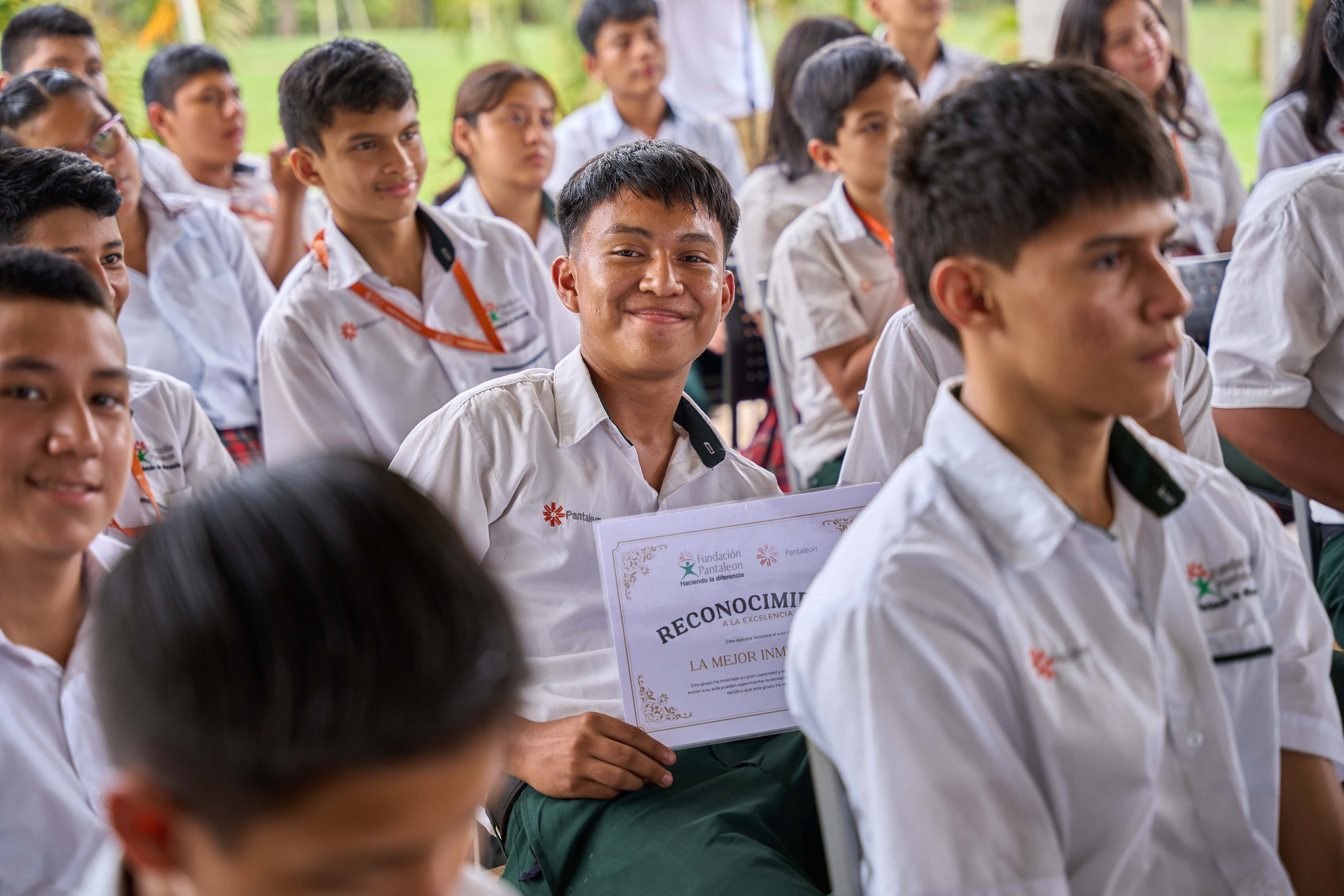 Estudiantes en uniforme blanco sentados en un evento, uno al centro sonríe sosteniendo un diploma de reconocimiento a la excelencia de Fundación Pantaleón.