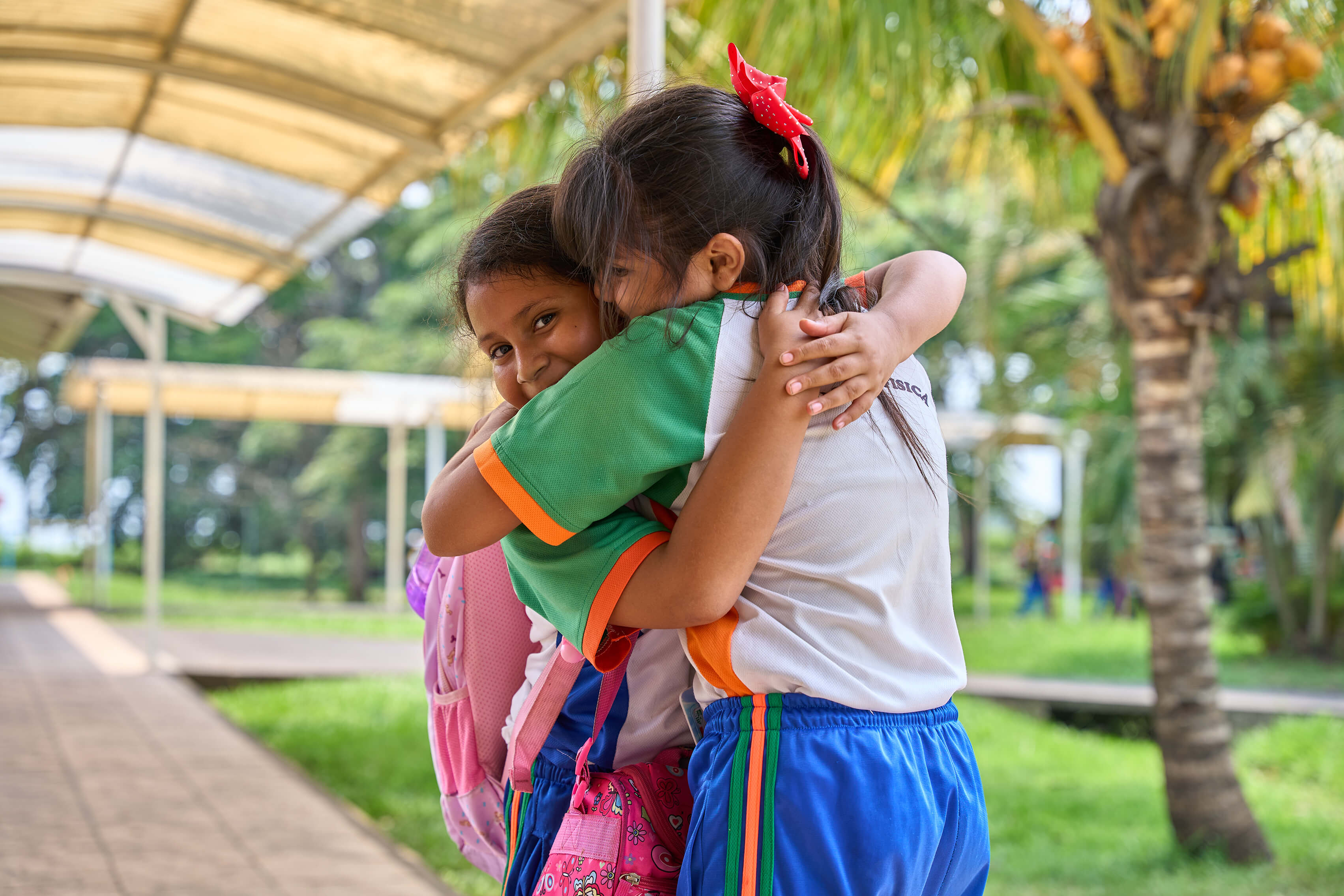 Dos niñas escolares abrazándose con sonrisas en un pasillo al aire libre con árboles y césped al fondo.
