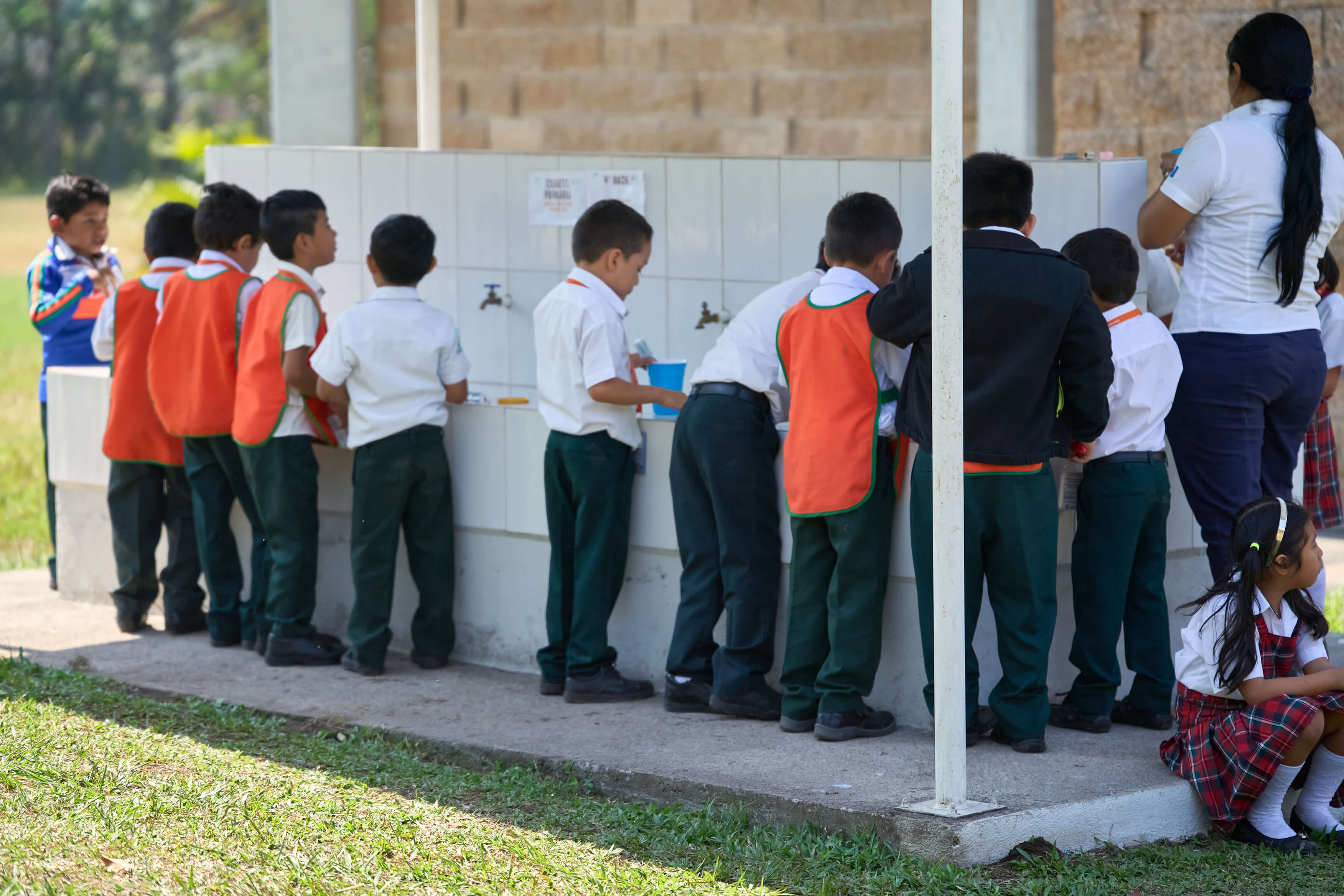 Niños y una mujer en un área al aire libre junto a un lavabo de azulejos blancos, algunos niños llevan uniformes con pantalones verdes y camisas blancas, y otros llevan mandiles naranjas; una niña está sentada en el borde del área, vestida con falda a cuadros y camiseta blanca.