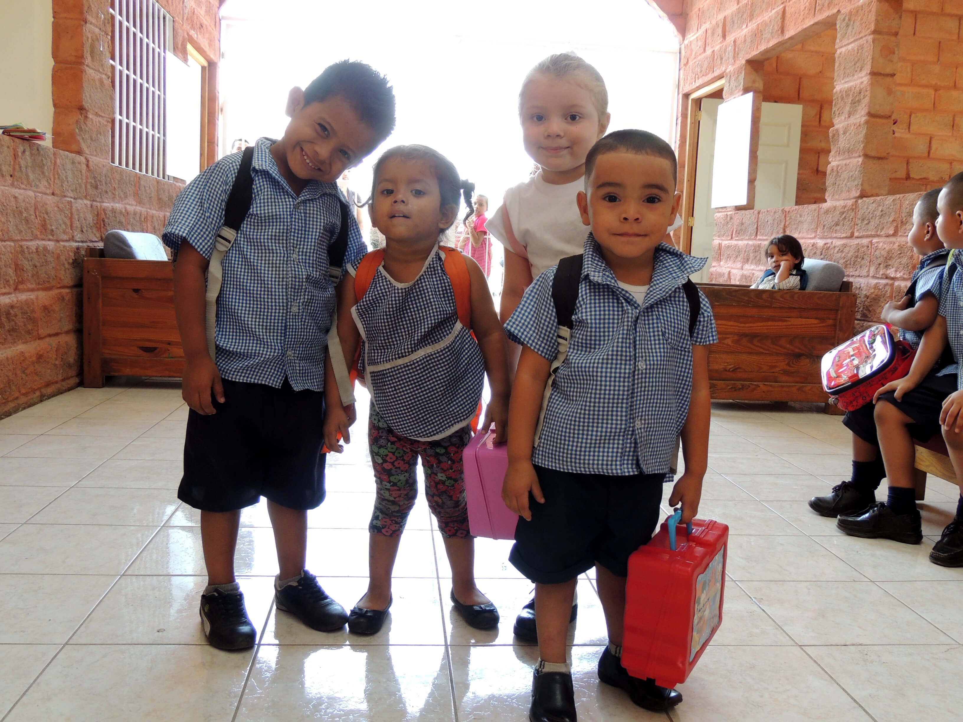 Cuatro niños pequeños en un pasillo escolar con paredes de ladrillo rojo y piso de baldosas claras; tres niños llevan uniformes escolares azules con camisa a cuadros y pantalones cortos con mochila y lonchera, y una niña con camiseta blanca y pantalones floreados sostiene una lonchera rosa.