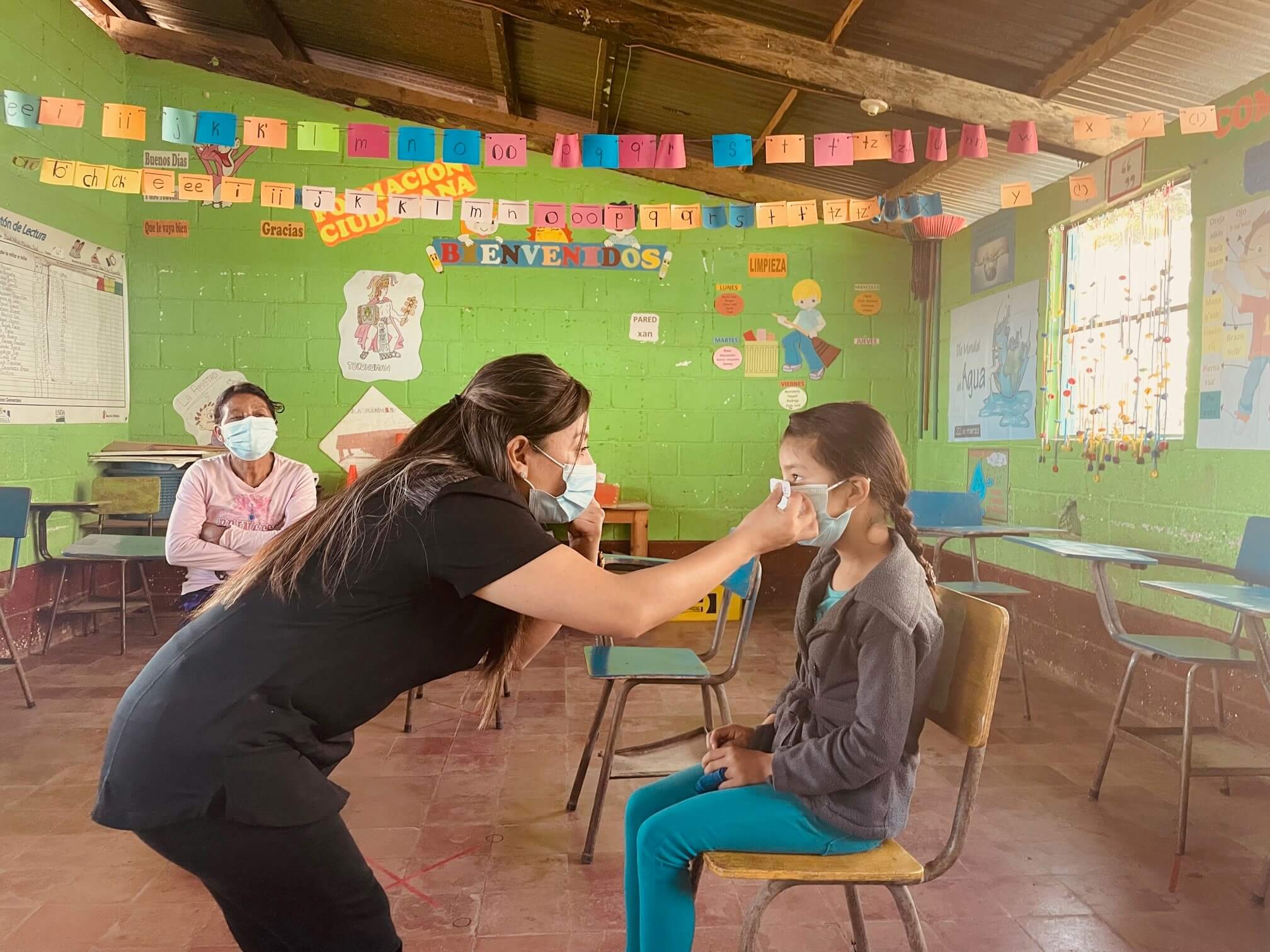 En un aula con paredes verdes decoradas con carteles educativos, una mujer cuida y ajusta el cubrebocas de una niña sentada en una silla, mientras otra persona observa sentada al fondo, todos usando mascarillas.