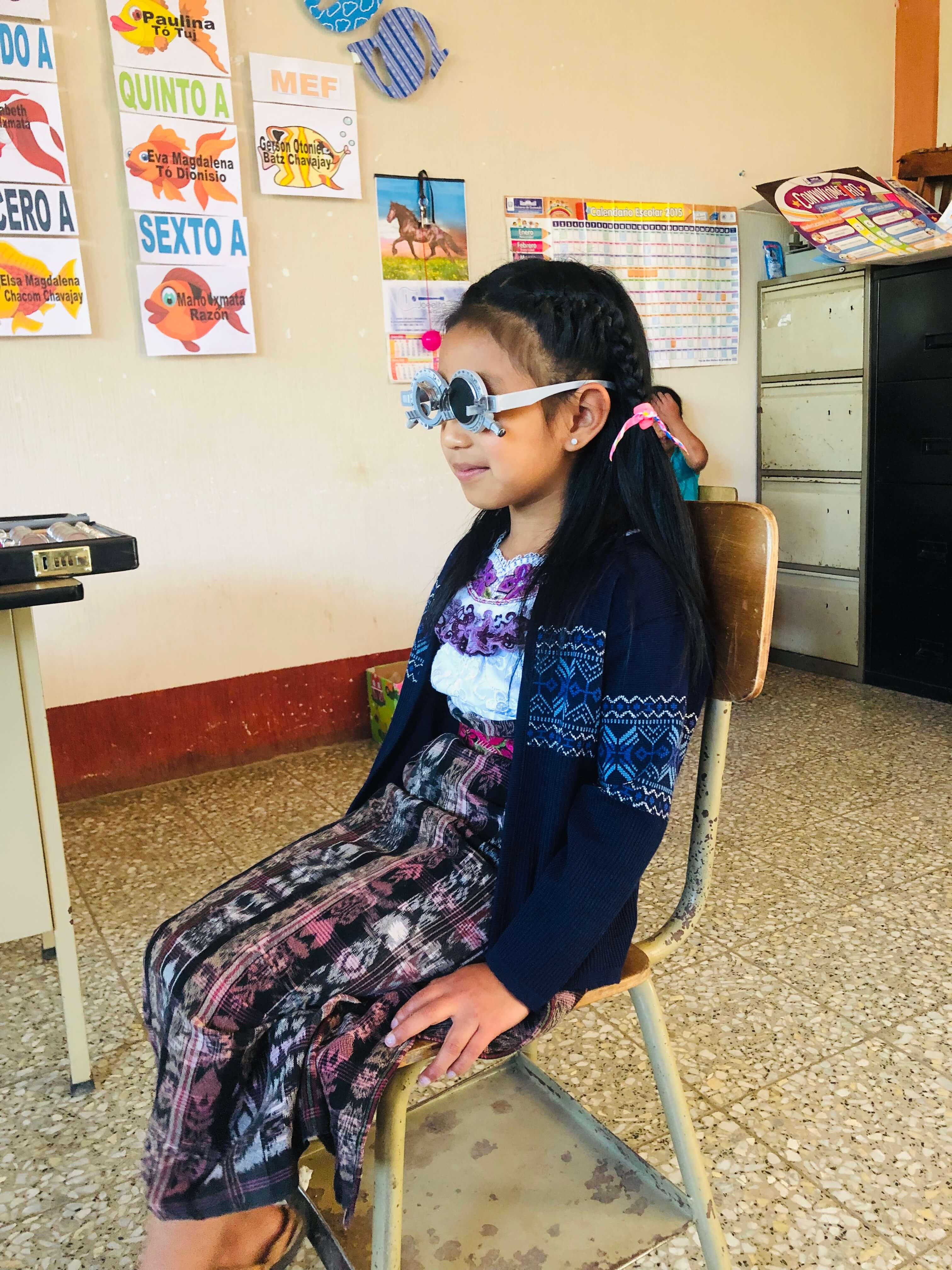 Niña maya con gafas de prueba óptica, vestida con ropa tradicional, sentada en una silla dentro de un aula con carteles y calendario en la pared.