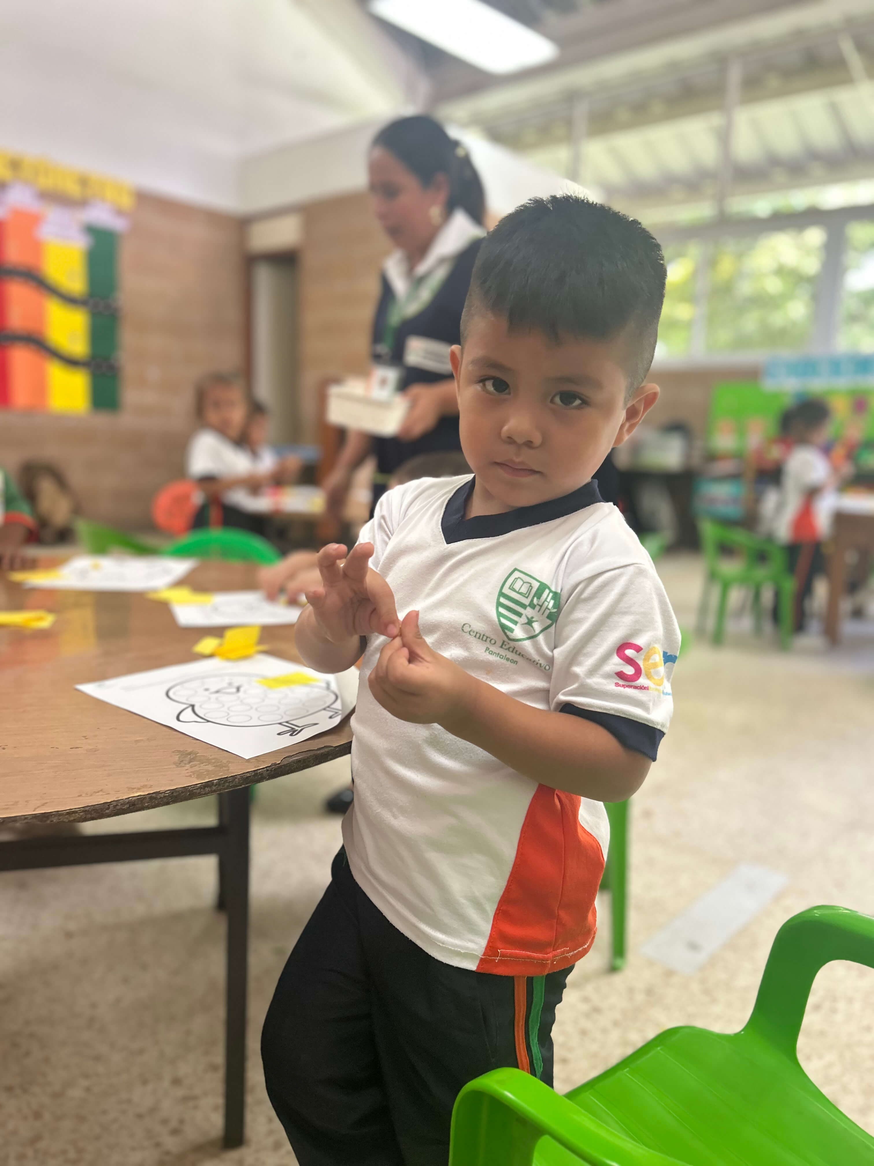 Niño pequeño en uniforme escolar blanco con detalles verdes y naranjas, de pie junto a una mesa con dibujos para colorear en un aula con sillas verdes y otros niños y una maestra al fondo.