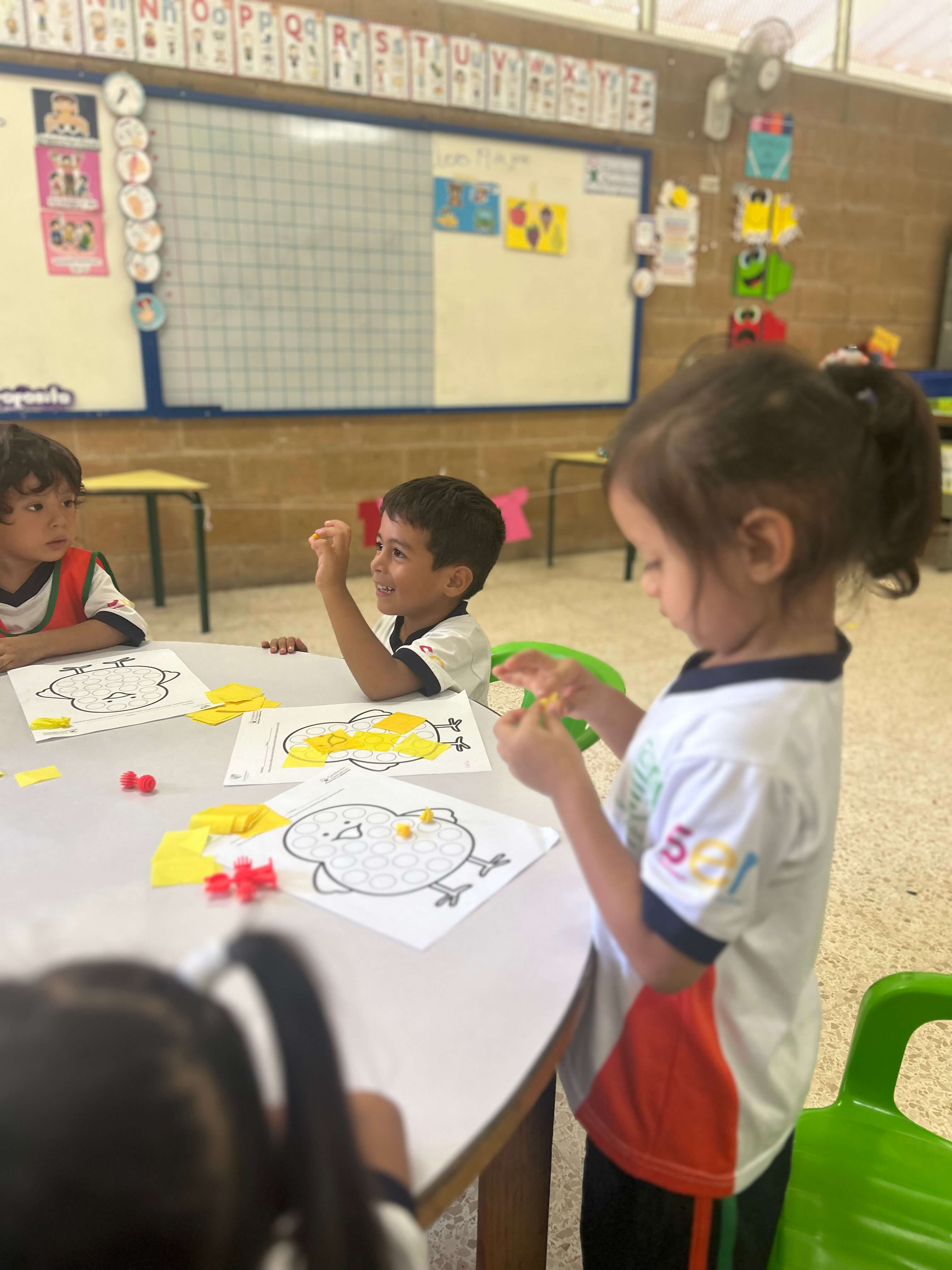 Niños pequeños en un salón de clases sentados alrededor de una mesa redonda realizando una actividad manual con papel amarillo y hojas con dibujos de pollitos, algunos niños están pegando los papeles mientras otros observan. En el fondo se ven carteles y un pizarrón con letras del alfabeto.