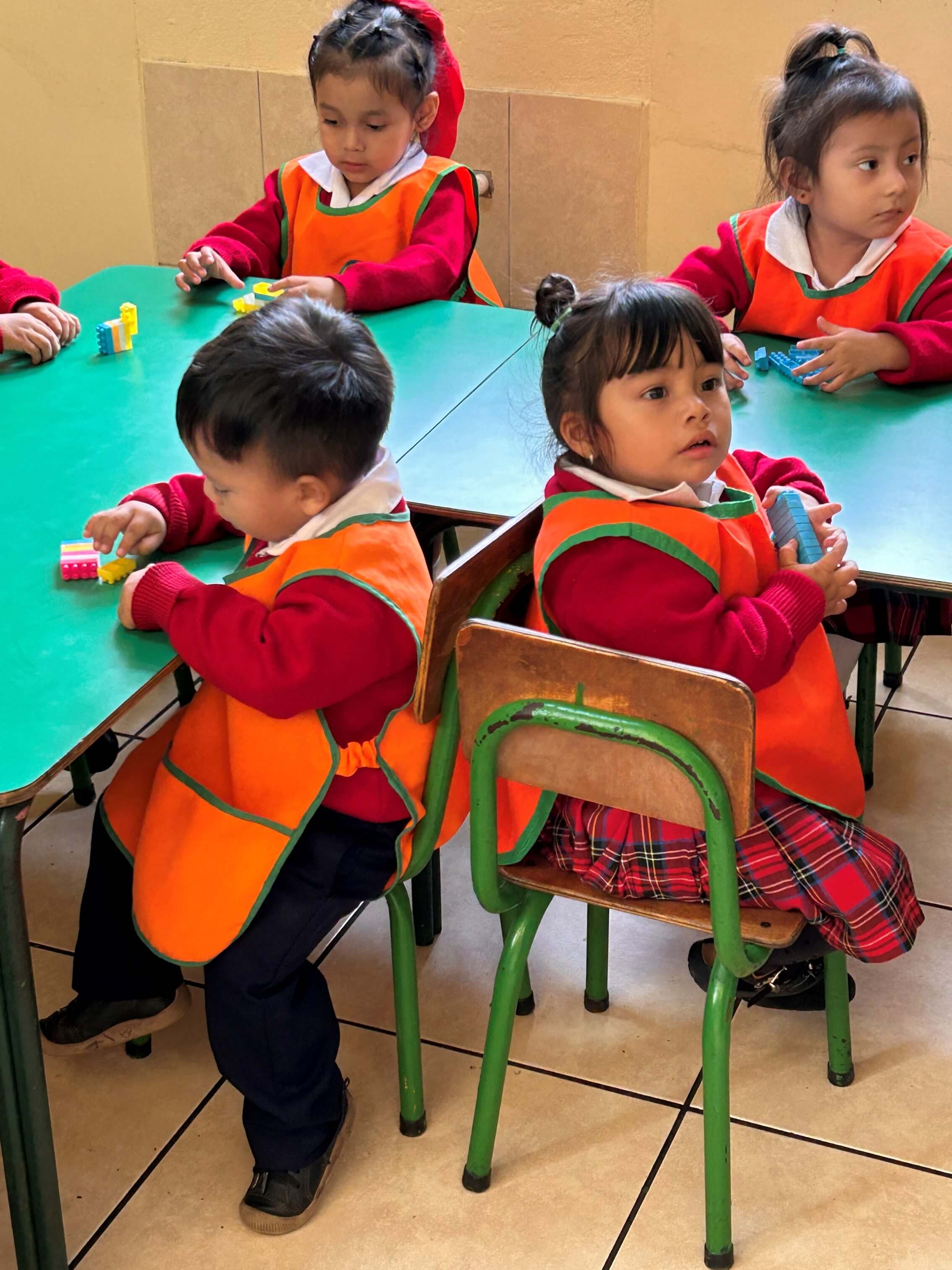 Cuatro niños pequeños en un aula sentados en sillas verdes y jugando con bloques de construcción de colores sobre mesas verdes, vestidos con uniformes que incluyen camisetas rojas, delantales naranjas y faldas o pantalones escolares.