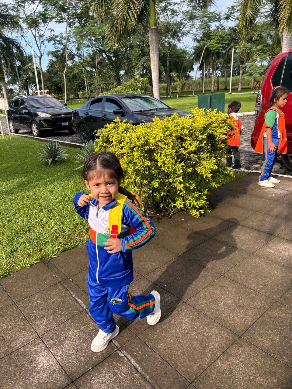 Niña pequeña con mochila amarilla y uniforme deportivo azul, blanco, verde y naranja, caminando y sonriendo en una acera junto a arbustos verdes, con otros niños y autos estacionados al fondo en un área con palmeras.