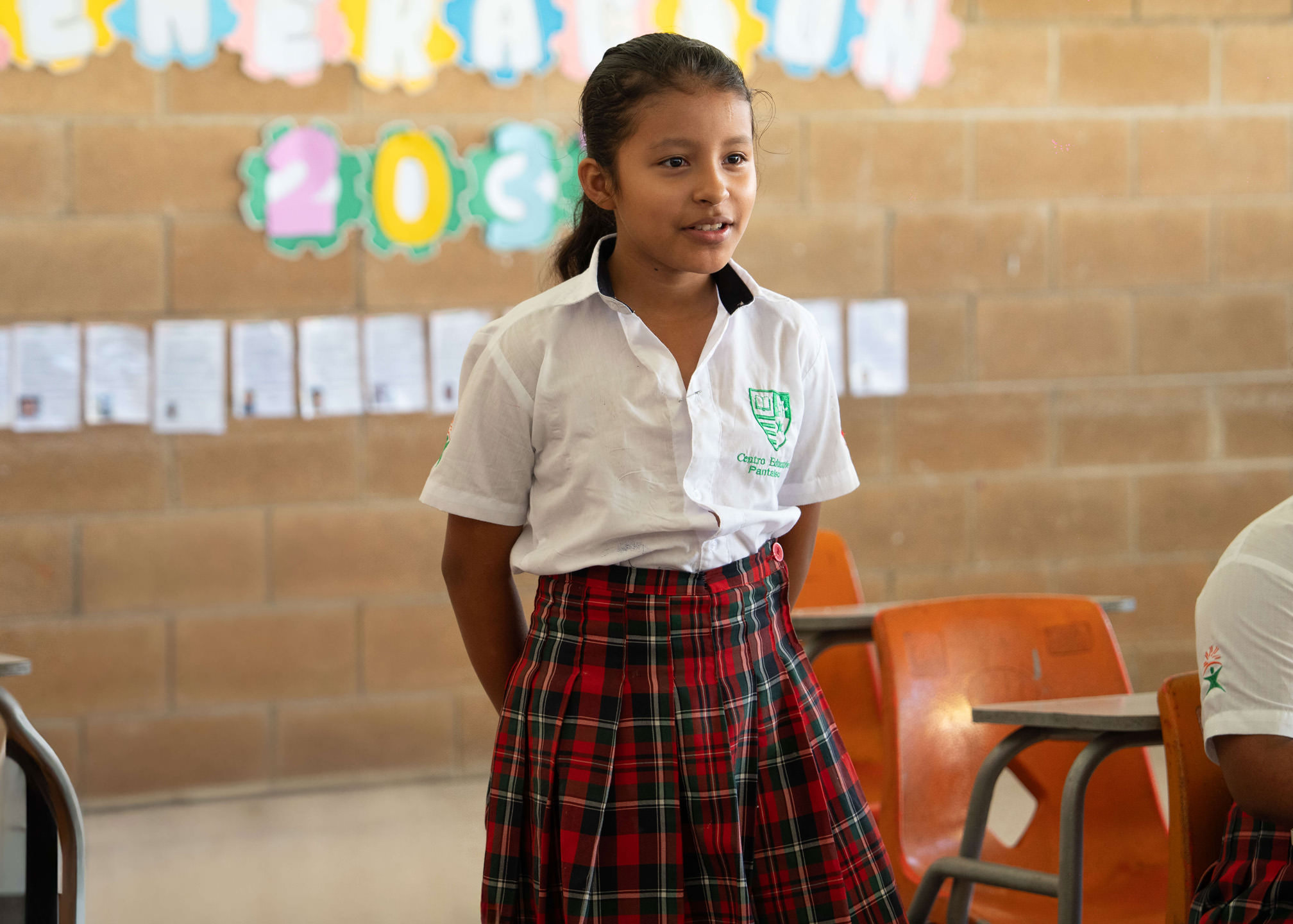 Niña con uniforme escolar blanco y falda de cuadros rojos y verdes de pie en un salón de clases con sillas naranjas y un muro de ladrillo con decoraciones coloridas y papeles pegados.