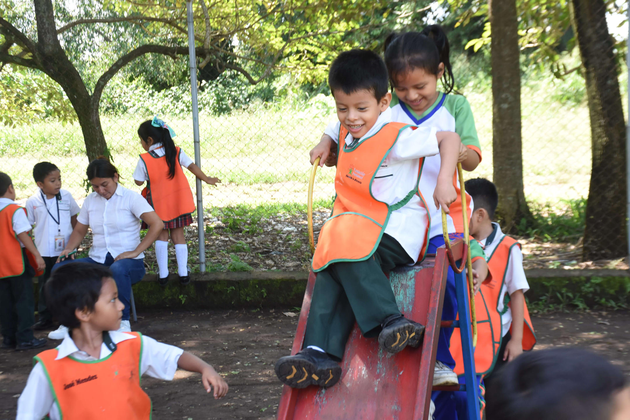Niños jugando en un parque escolar, dos niños se deslizan juntos por un tobogán rojo mientras otros niños y una maestra observan y juegan alrededor.