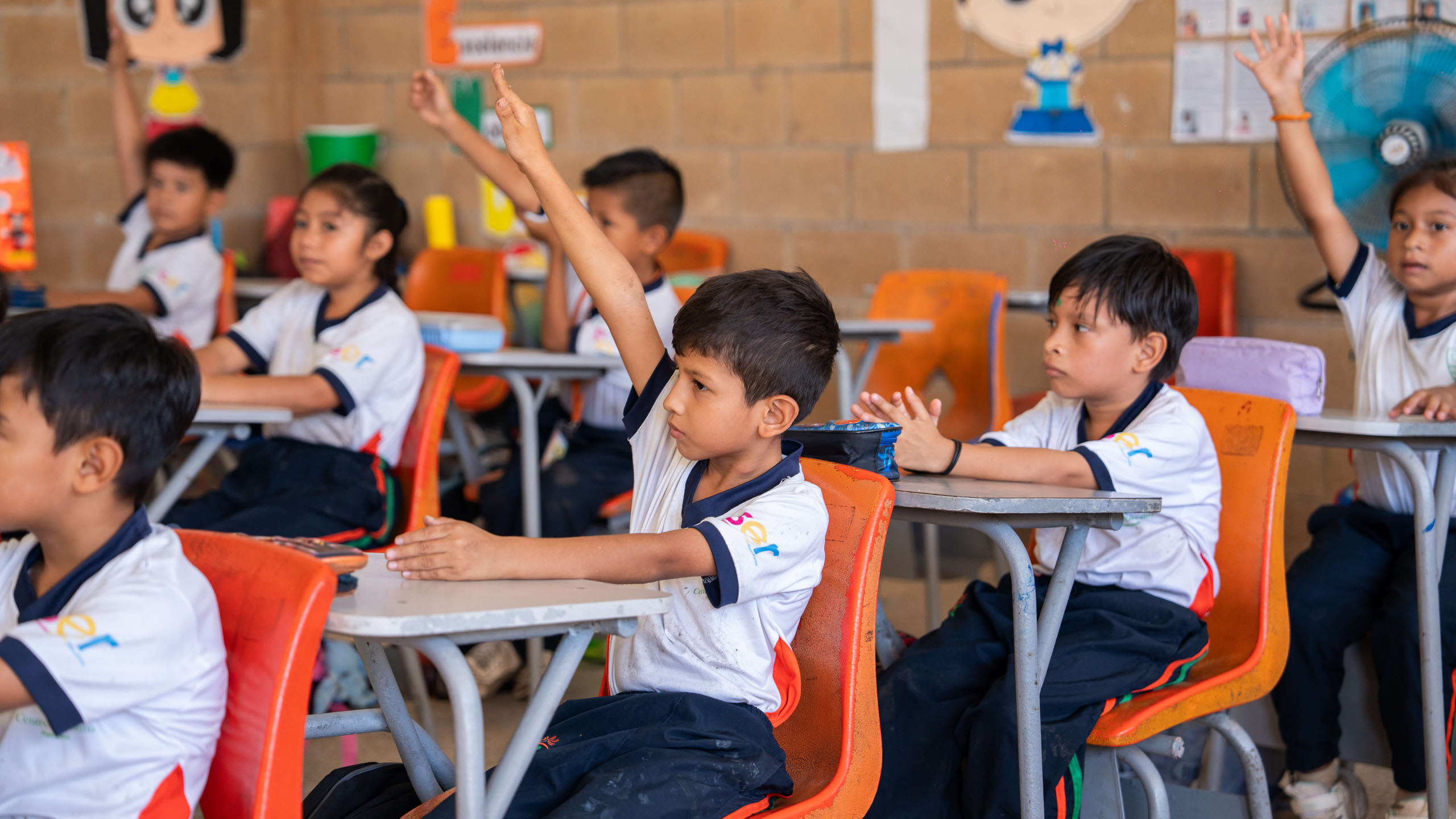 Niños en un aula escolar sentados en escritorios naranjas con uniformes blancos y pantalones oscuros, varios de ellos levantan la mano para participar.