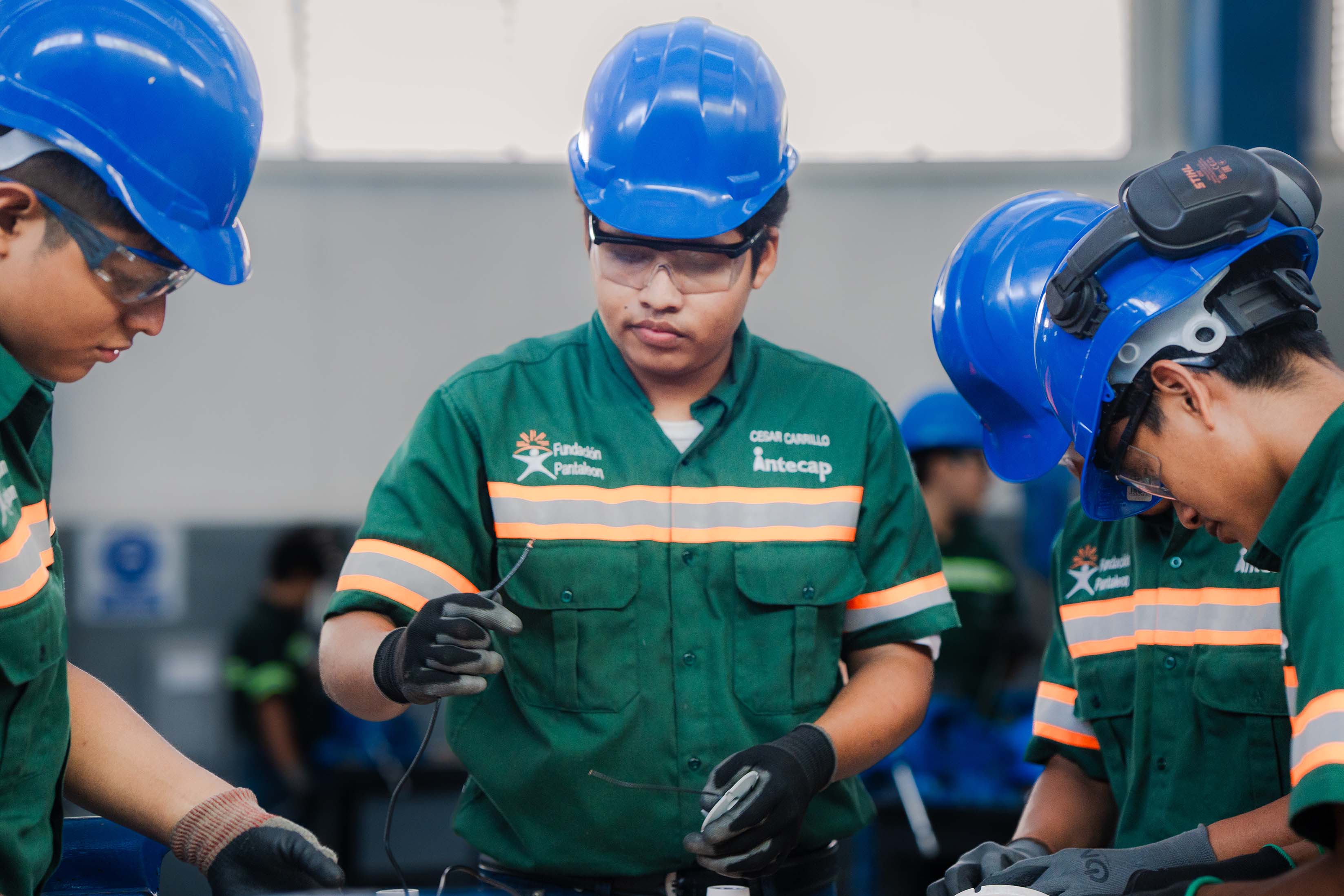 Cuatro hombres jóvenes con casco azul, gafas de seguridad y uniformes verdes con franjas naranjas trabajan juntos en un taller industrial, manipulando cables y herramientas.