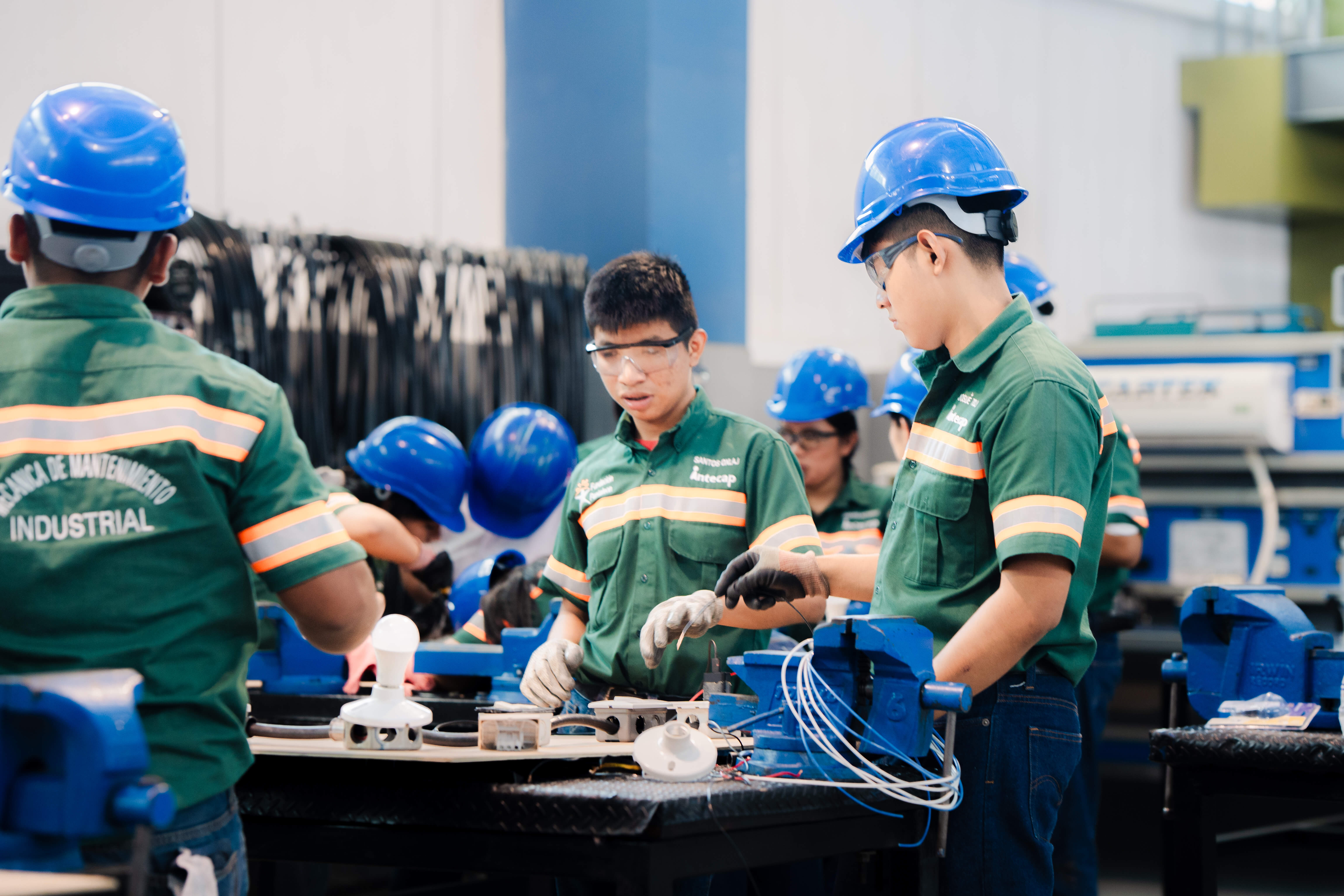 Estudiantes con uniformes verdes y cascos azules trabajan en un taller industrial, manipulando herramientas y componentes eléctricos sobre mesas de trabajo.